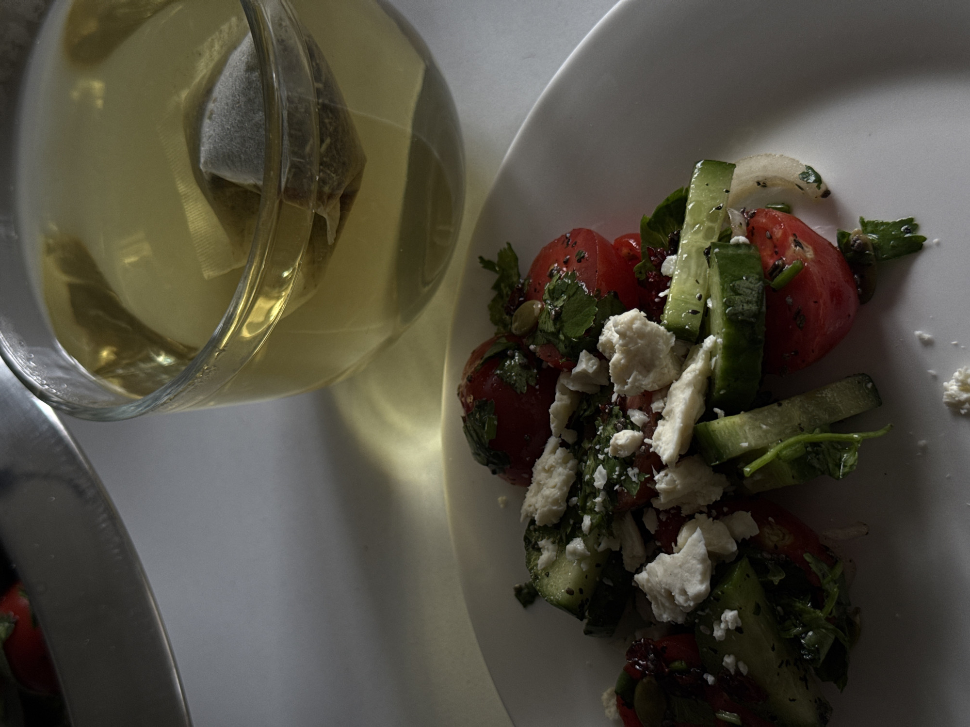 A high-angle shot depicts a white plate with a salad next to a glass teapot filled with tea. The salad consists of chopped tomatoes, cucumber, herbs, and crumbled white cheese. The tea in the teapot has a light yellow hue, with a teabag steeping inside. The scene is well-lit, casting shadows on the plate and the table, suggesting an indoor setting.