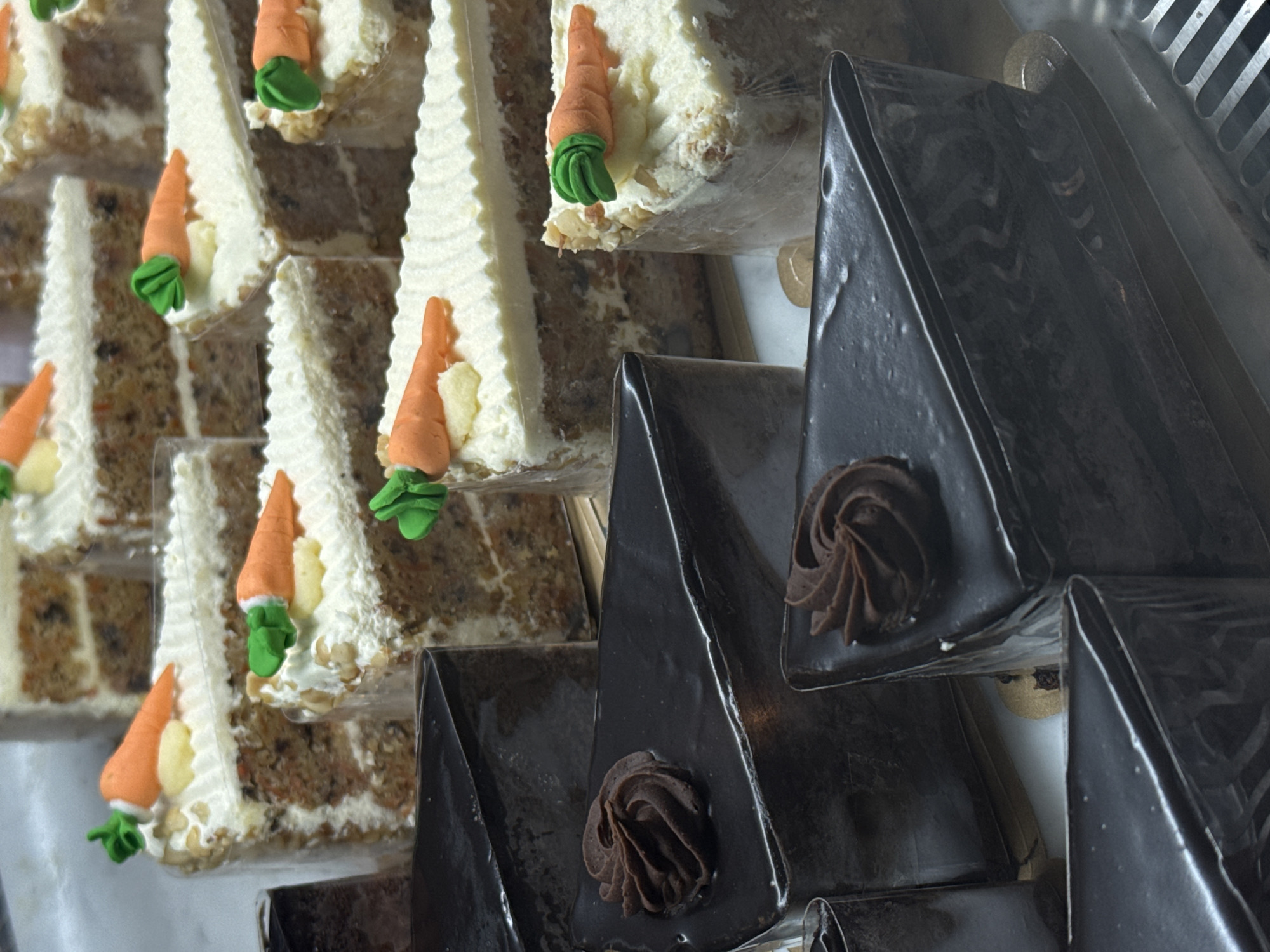 This image captures a close-up, high-angle view of various cake slices displayed, likely within a bakery or cafe setting in North Kuta, Indonesia. The display features two distinct types of cake, arranged in rows and individually packaged.
On the left and center, multiple slices of carrot cake are prominently featured. These triangular slices reveal layers of moist, light brown cake speckled with what appears to be carrots and possibly nuts. Each slice is generously frosted with a thick layer of white cream cheese frosting, applied with a decorative, ridged pattern along the edges. The carrot cake slices are adorned with small, intricate marzipan or fondant carrots, featuring orange bodies and green leafy tops. These slices are individually contained in clear plastic, suggesting they are ready for sale or serving.
To the right, several slices of chocolate cake are visible. These triangular slices are coated entirely in a dark, glossy chocolate glaze, giving them a sleek and decadent appearance. Each chocolate cake slice is topped with a single, neatly piped swirl of rich chocolate frosting, providing a textural contrast to the smooth glaze. Like the carrot cakes, these are also likely individually packaged or positioned in a way that implies individual portions.
The cakes are arranged on what appears to be a tiered or slanted display shelf, with some metallic elements of the display unit visible in the background, particularly on the far right. The overall lighting is bright and even, illuminating the cakes and their details. No people or visible text are present in the image.
