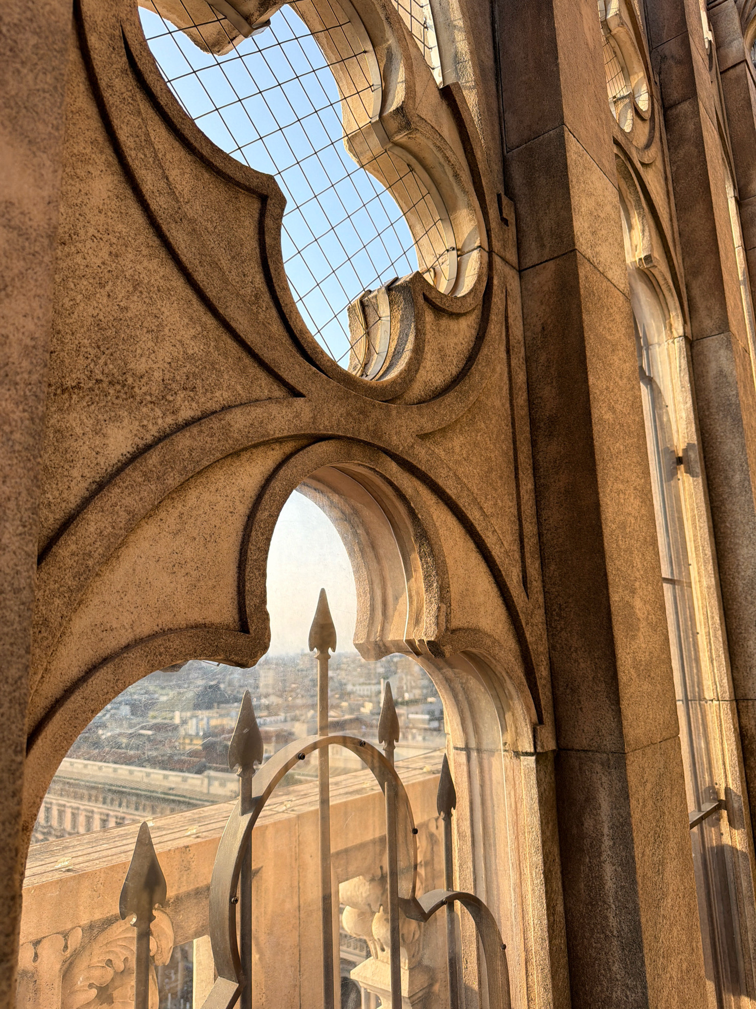 The image captures a view from the rooftop of the Duomo di Milano, Milan, Italy. The foreground features intricate stone architectural elements, characteristic of Gothic style, with arched openings and decorative carvings. Through an archway, a glimpse of the city of Milan is visible, with buildings and rooftops stretching into the distance. A metal railing with pointed finials runs along the edge of the rooftop, providing a sense of perspective. The lighting suggests it is either sunrise or sunset, casting warm golden hues on the stone and creating long shadows. The sky is clear and blue, visible through the patterned openings in the architecture.