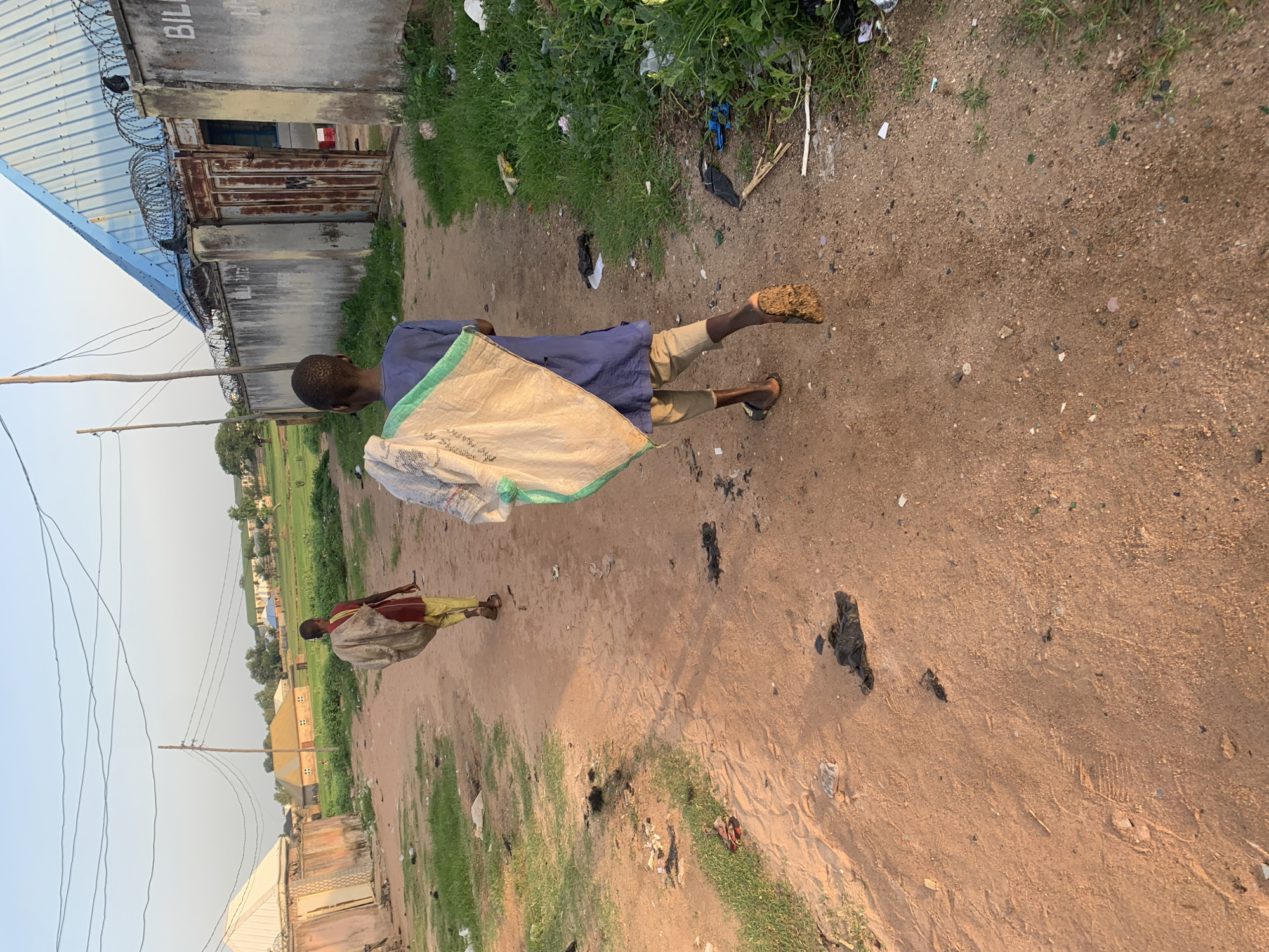 Two young boys are walking away from the camera on a dirt path in Jalingo, Nigeria. The boy in the foreground is wearing a blue tunic and tan trousers, carrying a large, light-colored sack over his right shoulder. The sack appears to be made of woven material and has a green trim along the top. The boy's left leg is extended forward, with his foot in a dark sandal that is partially covered in dirt. His right leg is behind him, with his heel lifted off the ground, showing the sole of his sandal also covered in dirt.
The second boy, further up the path and slightly to the left, is wearing a dark red tunic and yellow trousers. He is also carrying a large sack over his left shoulder. This sack appears to be a darker color, possibly brown or grey. His legs are visible, and he is also walking away from the camera.
The path is made of reddish-brown dirt and shows signs of foot traffic and scattered debris, including dark clumps of material and some white and green plastic. To the left of the path are patches of green grass with some scattered litter. Further in the background on the left, a building with a white roof and a light-colored wall is visible.
To the right of the path, there is more green grass and dense vegetation. In the background, several buildings can be seen. One building is a light grey structure with a rusted metal gate and a bright blue corrugated metal roof. This building is topped with barbed wire. The word "BILL" is visible in black capital letters on the wall of this building. Other buildings with various roof colors, including green and brown, are visible further in the distance, suggesting a more urban or semi-urban environment. Overhead, a network of electrical wires is strung between poles. The sky is clear and appears to be late afternoon or early evening, with soft lighting casting shadows.