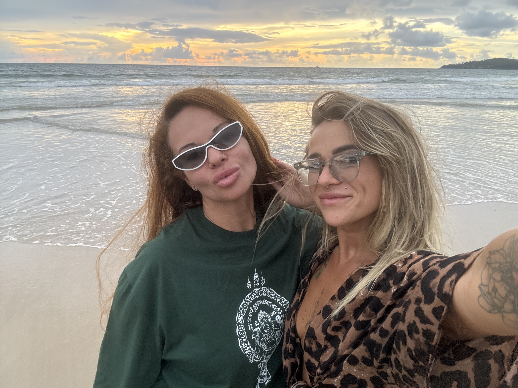Two women are taking a selfie on a beach at sunset in Choeng Thale, Thailand. The woman on the left has long auburn hair, is wearing white-framed sunglasses and a dark green t-shirt with a white circular design on it. The woman on the right has blonde hair, is wearing glasses and a leopard-print top, and has tattoos on her arm. The ocean is behind them, with gentle waves and the sky is filled with colorful clouds.