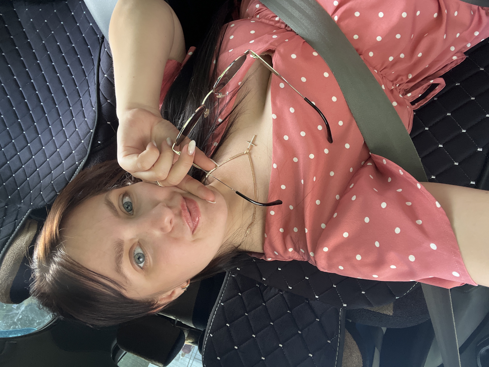A woman is in a car, wearing a pink polka dot dress and a seatbelt. She holds a pair of sunglasses. A gold necklace with a cross pendant is visible. The car has black quilted seat covers. The location is Chaikovsky, Russia.