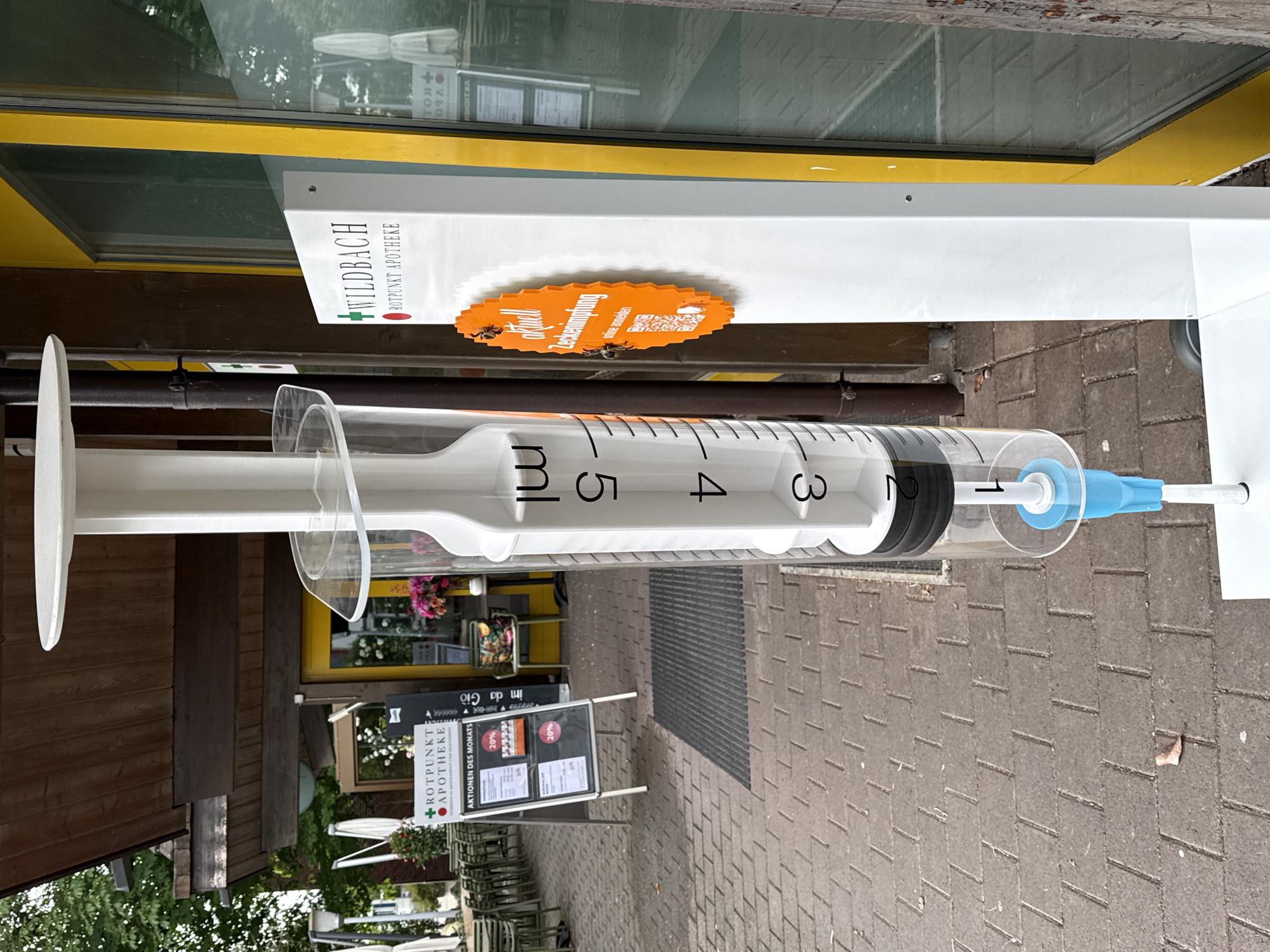 An oversized replica of a medical syringe stands upright in front of a building in Embrach, Switzerland, serving as a prominent display. The syringe features a white plunger, a clear barrel marked in milliliters (mL) with numbers from 1 to 5, a black rubber stopper, and a light blue cap covering the needle end.
Behind the syringe, a white rectangular sign for "WILDBACH ROTPUNKT APOTHEKE" is visible, featuring a green cross and a red circle logo. Attached to this stand is an orange oval sign with jagged edges, reading "aktuell Zeckenimpfung" (current tick vaccination) and "online anmelden" (register online), accompanied by a QR code, indicating a health campaign.
The scene appears to be an outdoor pedestrian area or street. To the left, a wooden building facade is visible, and further back, another storefront with a black sandwich board sign reads "+ROTPUNKT APOTHEKE," "AKTIONEN DES MONATS" (promotions of the month), and "im da Giò," possibly the name of a restaurant or shop, offering "20%" discounts. Small tables and chairs suggest an outdoor seating area for "im da Giò."
The ground is paved with rectangular bricks. Reflections in the building windows show parts of the street and possibly other buildings. The lighting suggests it is daytime, likely an overcast day as there are no strong, distinct shadows. No people are present in the image, making it a static scene focused on the commercial displays.