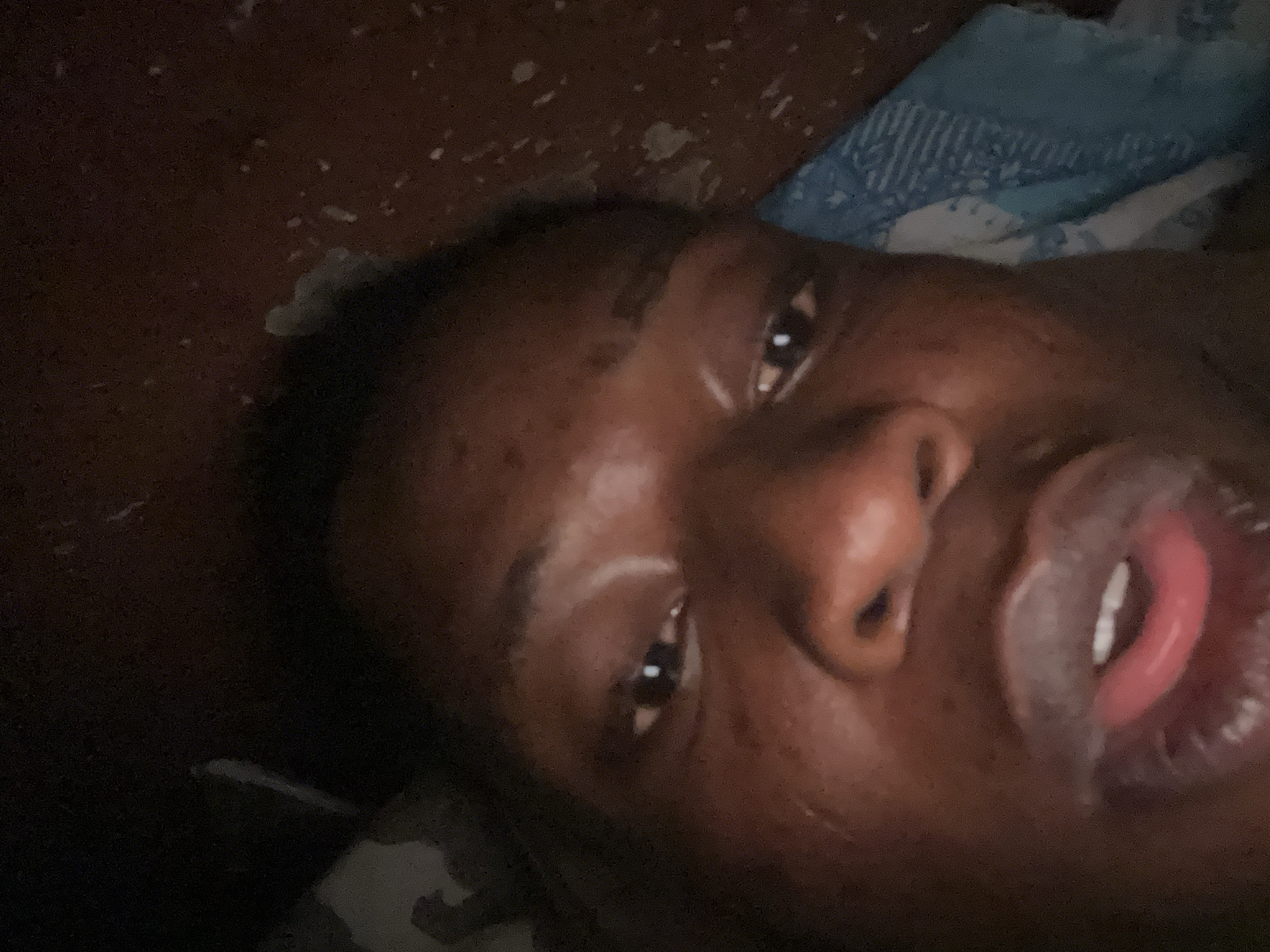 Close-up selfie of a Black person lying down. The person's face is angled towards the camera. The person has brown eyes, a wide nose, and open lips revealing their tongue. There is a blue and white patterned textile visible in the background. The environment is dark and difficult to discern. The location is unknown.