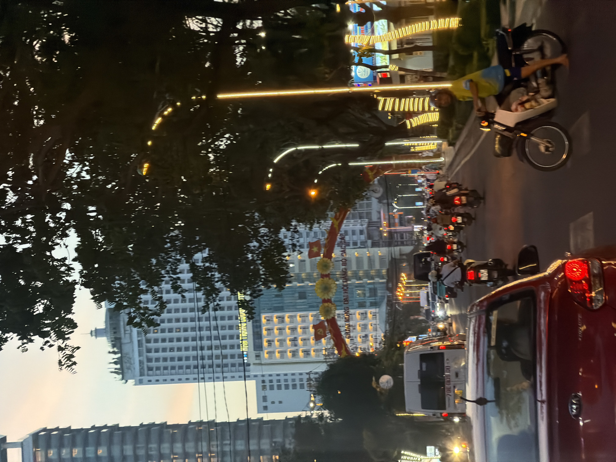 The image depicts a street scene in Nha Trang Ward, Vietnam, at dusk. Several buildings are visible, including tall, modern high-rises with illuminated windows and decorative lights. Trees line the street, some adorned with strings of yellow lights, creating a festive atmosphere. The road is busy with traffic, primarily motorcycles, and a few cars and a van. People on motorcycles are seen wearing helmets and casual clothing. One motorcyclist in the foreground is wearing a yellow and blue shirt and dark shorts, with bare feet. The sky is a soft orange and gray, indicating the setting sun. An archway decorated with red banners and flags is visible in the mid-ground. The overall impression is a lively urban environment during evening hours.