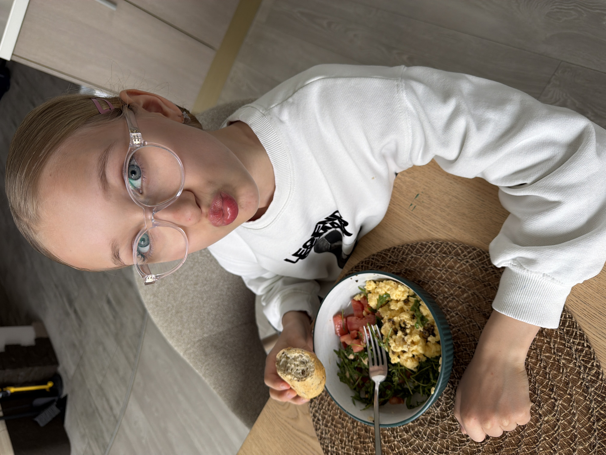 A young girl with blonde hair pulled back and wearing round glasses is sitting at a table in an interior setting, possibly a residential space in Dreiliņi, Latvia. She is wearing a white sweatshirt and holding a piece of bread in one hand. In front of her is a bowl containing scrambled eggs, diced tomatoes, and green leafy vegetables. A fork is placed in the bowl. The table is covered with a woven placemat. The floor and chair near the table are visible in the background.