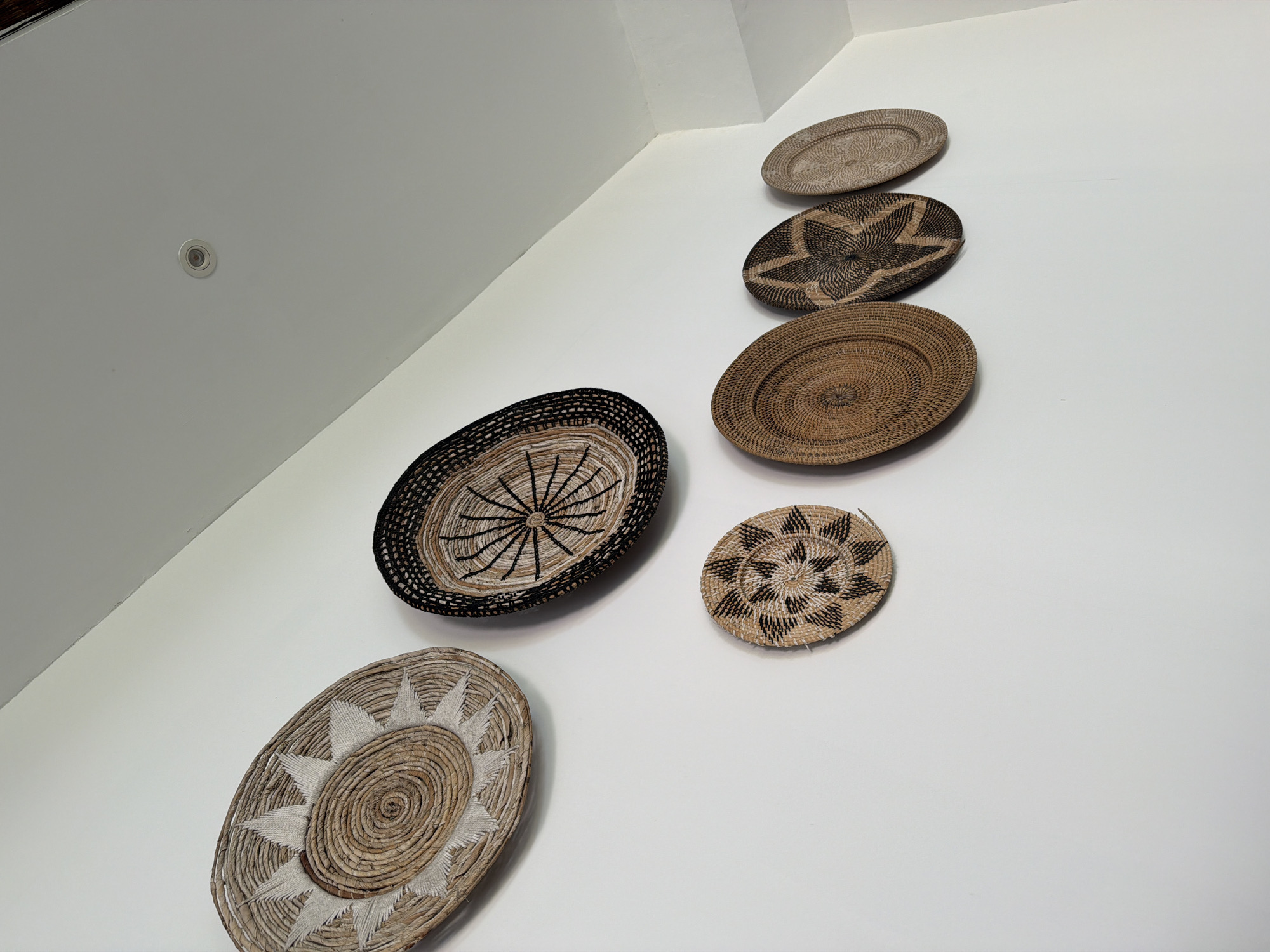 This media file captures an indoor scene featuring a white wall and ceiling adorned with six decorative woven baskets of varying sizes and patterns. The baskets, primarily round or oval, are hung on the wall in a diagonal arrangement, ascending from the bottom left towards the top right of the frame.
The individual baskets showcase intricate craftsmanship with different natural fiber colors, ranging from light beige to dark brown and black. Their designs include spiral patterns, radial lines, and star-like motifs. For example, one large oval basket in the lower left features a central spiral with radiating white elements, while another larger oval basket higher up displays a distinct dark brown star pattern against a lighter background. A smaller, circular basket near the center right exhibits a dark patterned design.
The wall and ceiling are a uniform white, creating a clean and minimalist backdrop for the rustic-looking woven art. A single recessed light fixture is visible on the white ceiling in the upper left corner. The image focuses primarily on the decorative elements, with no people or specific activity depicted, suggesting the baskets are intended as permanent wall decor. The light appears to be even, indicating an indoor setting with either artificial illumination or diffused natural light. Given the context of Kuta, Indonesia, these handcrafted baskets likely represent local artisan work, complementing a contemporary interior design aesthetic. There is no visible text in the image.