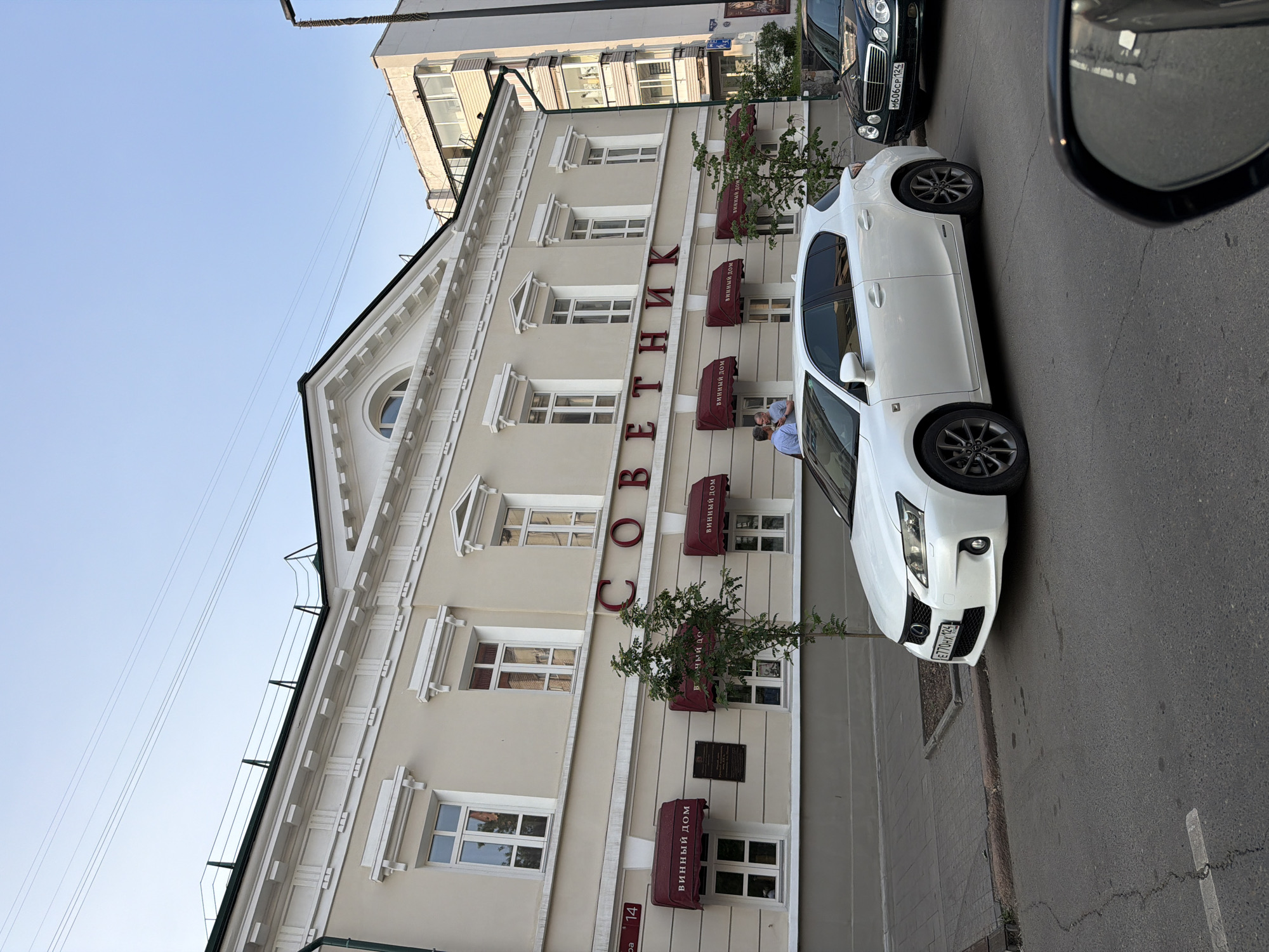 The image depicts a street scene in Krasnoyarsk, Russia. A light-colored, modern building with ornate architectural details lines the street. Red awnings with Cyrillic lettering that reads "ВИННЫЙ ДОМ" (Vinnyy Dom - Wine House) are visible above several windows on the ground floor. Large, red Cyrillic letters spelling "СОВЕТНИК" (Sovetnik - Advisor) are prominently displayed on the building's facade. Two individuals are seen standing and interacting on the sidewalk in front of the building.
Parked on the street are two cars: a white Lexus hatchback in the foreground and a dark-colored Mercedes-Benz sedan behind it. The white Lexus has a license plate that reads "E770HX 124". The Mercedes-Benz has a license plate that reads "M606CP 124". A street lamp is visible on the right side of the frame, and power lines crisscross the sky. The sky is clear and blue, suggesting it is daytime. The overall environment suggests a commercial area within the city.