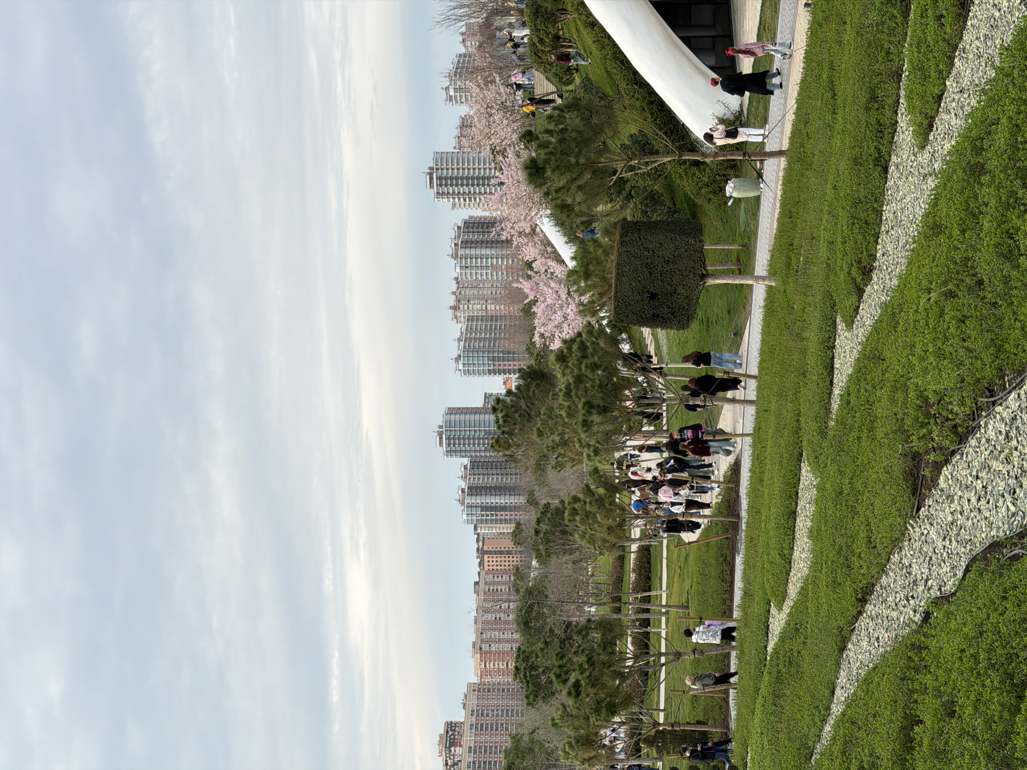 The image captures a park scene with modern architecture and landscaping. Tall residential buildings line the horizon under a cloudy sky. People stroll along a pathway lined with trees and manicured greenery. The ground is covered with grass and geometric patterns of white stones. Some people are sitting on benches and relaxing in the park. A portion of a modern white structure is visible on the right side. The location is likely the city of Valencia, Spain.