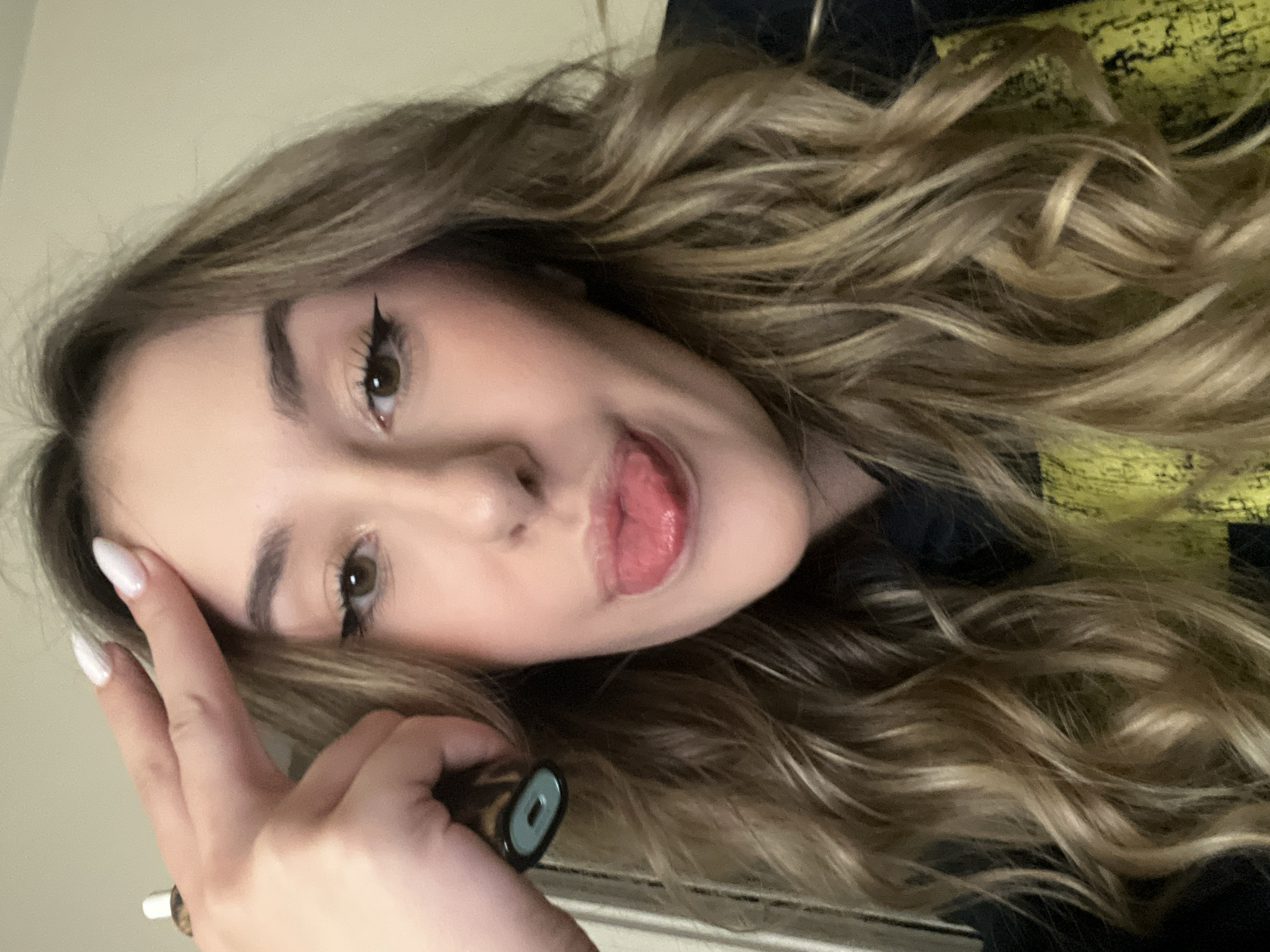 A young woman with long, wavy blonde hair is seen in a close-up, rotated perspective. She has fair skin, brown eyes, winged eyeliner, and is sticking her tongue out slightly. Her left hand is raised near her forehead, with white-tipped nails visible. In her hand, she holds a dark-colored object with a circular opening at one end. She is wearing a black hoodie with a yellow pattern on it. The background is a plain, light-colored wall. The location is not discernible and cannot be determined to be in Sofia, Bulgaria.