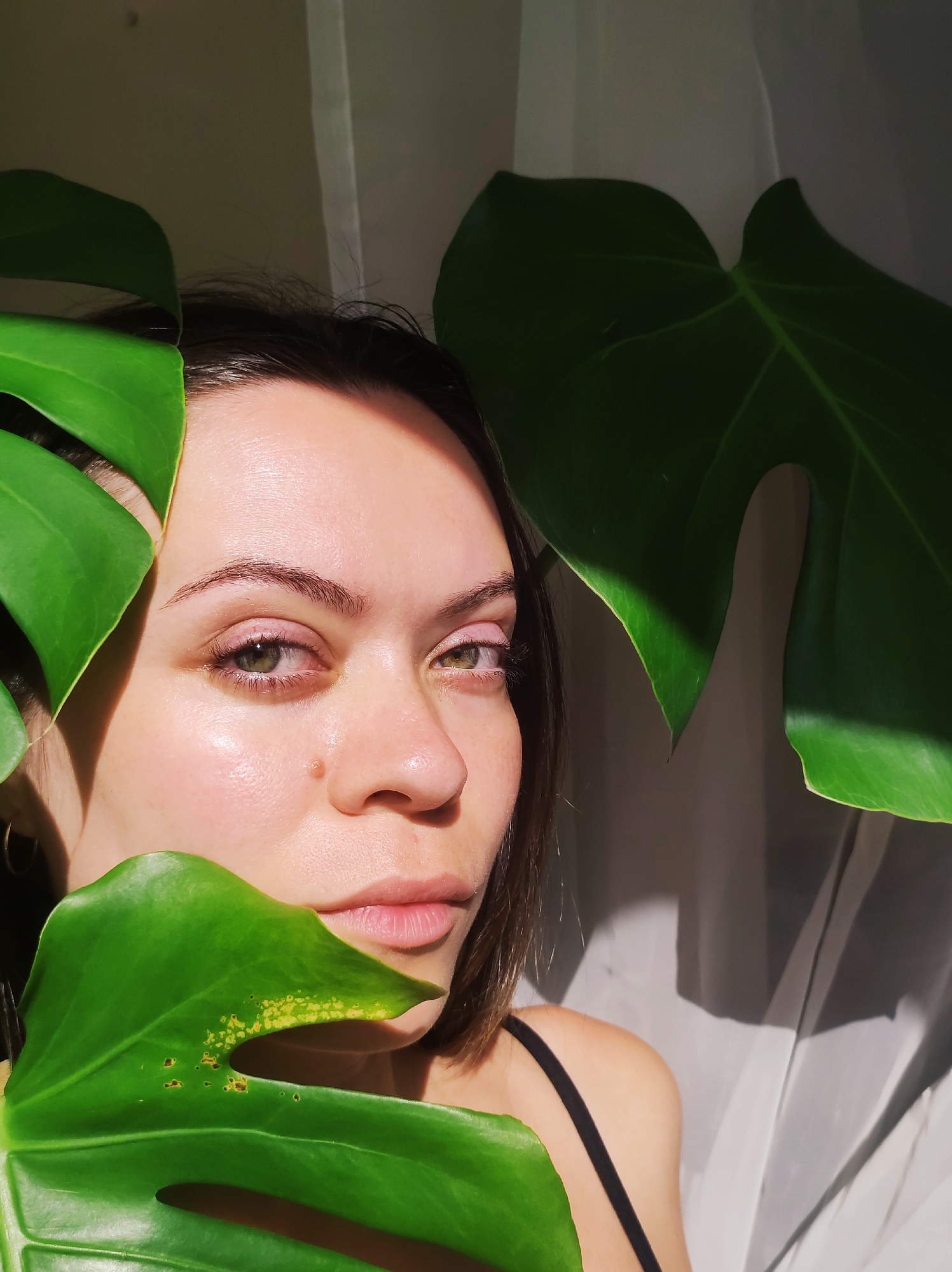 A young woman poses for a close-up photo in front of a white curtain, partially obscured by the large green leaves of a Monstera plant. The leaves are positioned on either side of her face and partially in front of her, with one leaf exhibiting some yellowed spots. The woman has fair skin, dark hair, and green eyes, and wears light pink eye shadow. She has a mole on her cheek near her nose. Her expression is neutral. A black strap of a garment is visible on her shoulder. The backdrop consists of a slightly wrinkled, light-colored fabric, likely a curtain, which casts shadows from the sunlight. The photo's location in the city of Chaikovsky, Russia cannot be determined from the visual information.