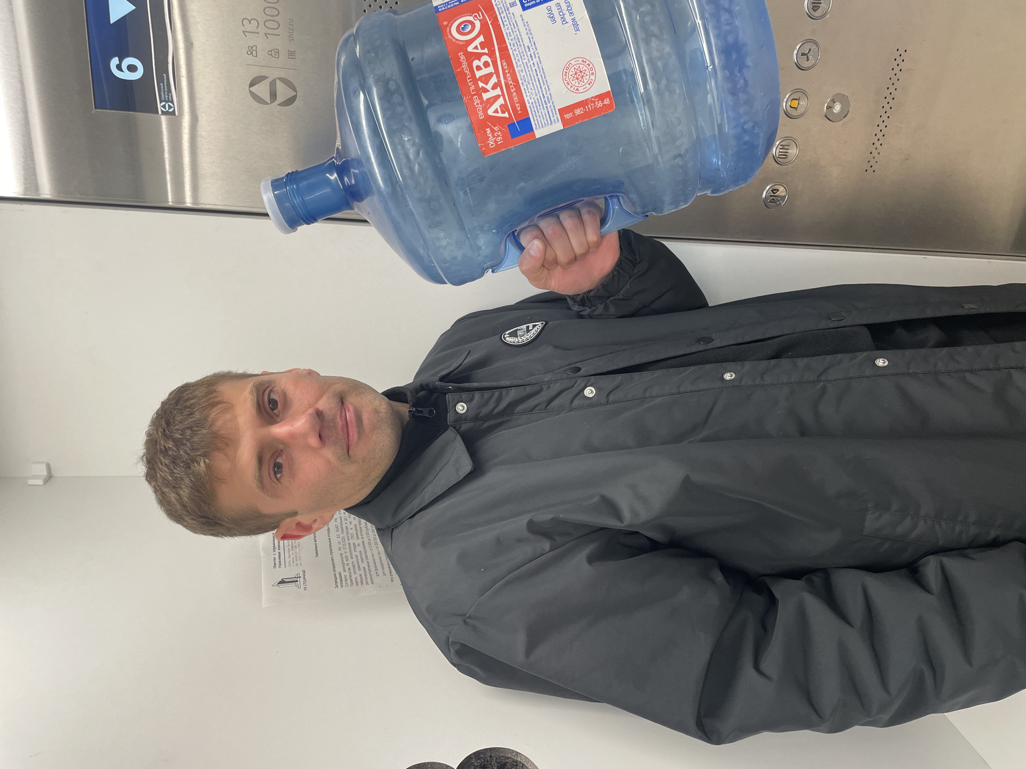 A man stands inside an elevator holding a large blue plastic bottle of water labeled "AKBAO" with Russian text. The elevator panel behind him shows buttons and the number "6". A sign is visible on the wall to the left of the man, with printed text in Russian, including "UK STOLICHNAYA." The sign references fines related to storing construction materials on public land. The location is Izhevsk, Russia, based on the water bottle brand and text on the sign. The man is wearing a black jacket.