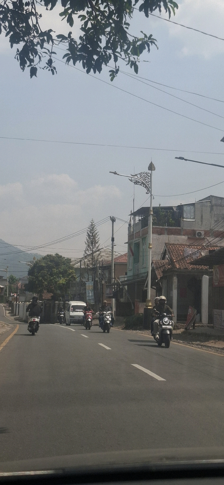 The image captures a street scene in Cilaku, Indonesia. The perspective is from a moving vehicle, looking forward along an asphalt road. Several motorcycles are visible, some with riders. In the foreground, a dark-colored motorcycle with two occupants is in the right lane. Further ahead, a white van is in the left lane, with multiple motorcycles on either side and behind it. The road is marked with white dashed lines. To the right of the road, there are buildings with tiled roofs, some appearing residential or commercial, and a decorative street lamp. Trees, including a tall evergreen, are present along the roadside and in the background. Utility poles and overhead wires are also a prominent feature. The sky is clear with some scattered clouds, and hills are visible in the far distance. The overall impression is of a typical, moderately busy day on a road in this Indonesian town.