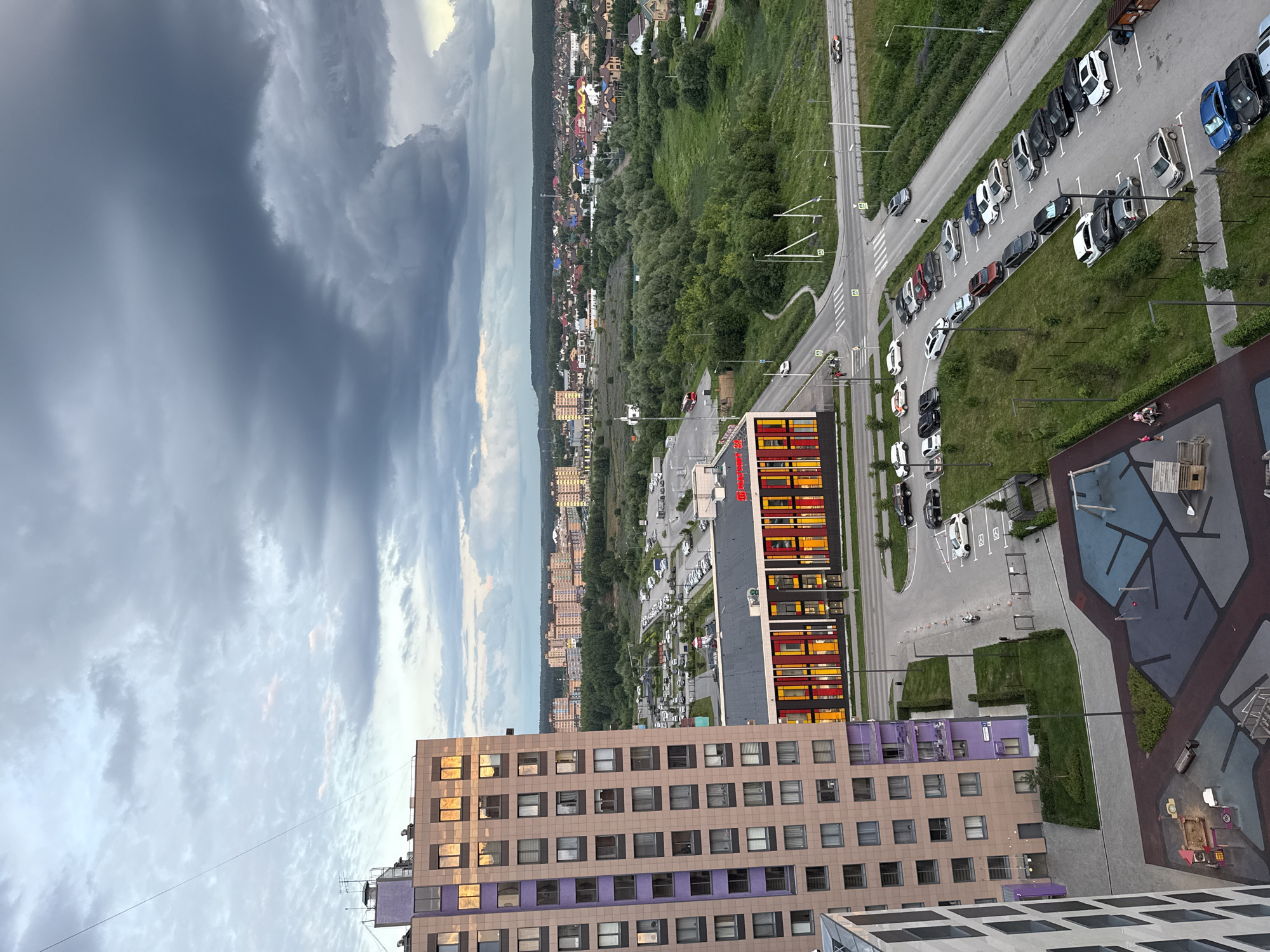 The aerial, eye-level shot captures an urban landscape in Perm, Russia, under a dramatic, overcast sky with heavy, dark clouds dominating the upper portion of the frame. In the foreground, a modern, multi-story apartment building with beige and purple facades stands prominently on the left. Adjacent to it, a building with a colorful facade featuring red, yellow, and orange vertical accents is visible.
A parking lot filled with various cars, including sedans and SUVs, is situated in the middle ground. A red bus is parked near a green grassy area. The scene extends to a residential area with numerous low-rise buildings and houses with pitched roofs, interspersed with dense green trees. A road with white lines and traffic signs traverses the middle of the image, with several cars driving on it. Two individuals are walking on a sidewalk near the road. A playground with swings, a sandbox, and a small wooden play structure is visible in the lower left corner. The overall environment appears to be a developing urban or suburban area.