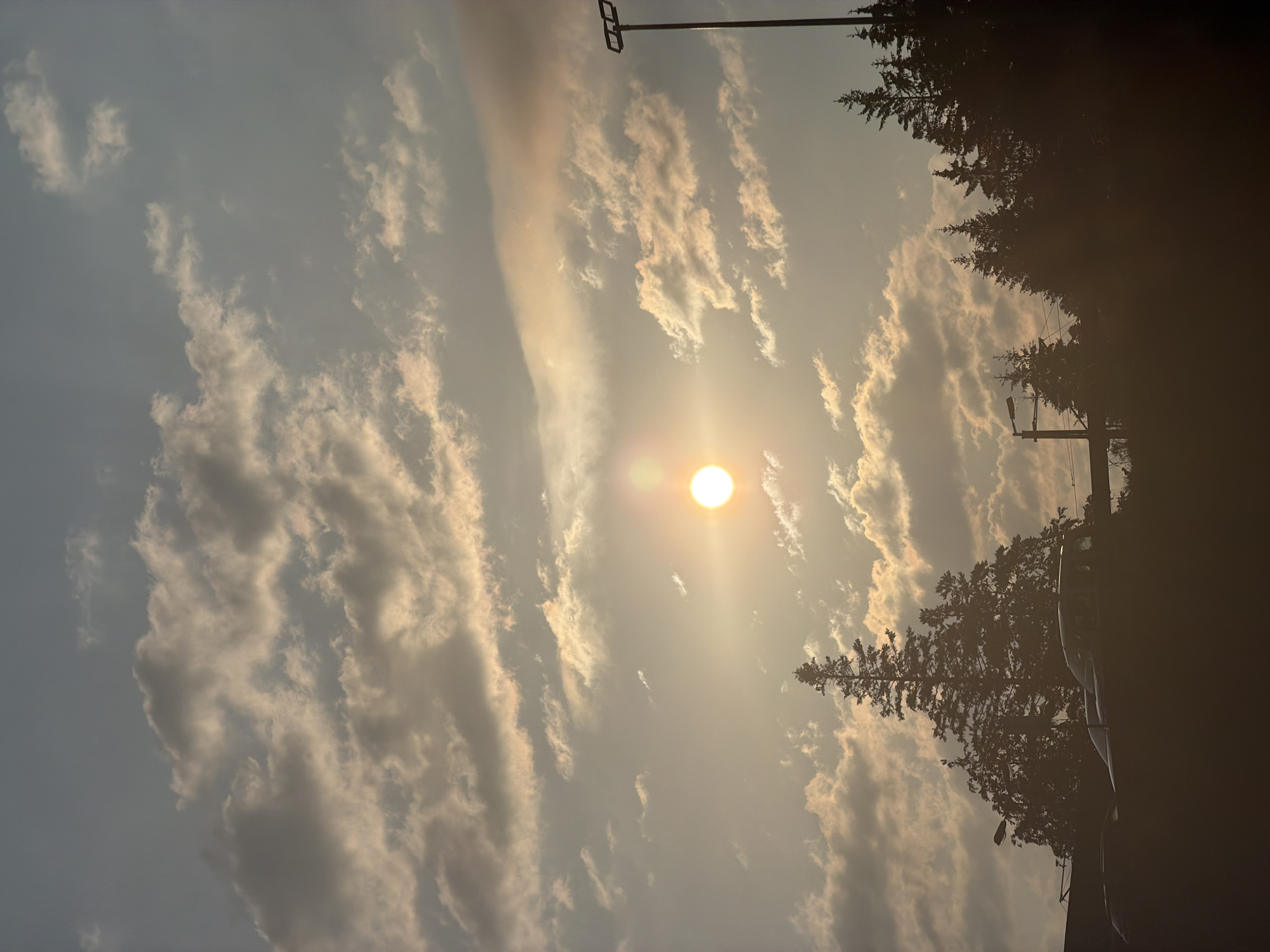 This image captures a sunset over a silhouetted landscape in Grainau, Germany. The sky is filled with scattered clouds, illuminated by the warm glow of the setting sun, which appears as a bright, round disc slightly off-center to the left. In the foreground, dark silhouettes of evergreen trees and what appear to be cars are visible, suggesting an urban or suburban edge. A tall lamppost with multiple lights stands on the right side of the frame, also silhouetted against the bright sky. The overall impression is one of a calm, late afternoon or early evening scene.