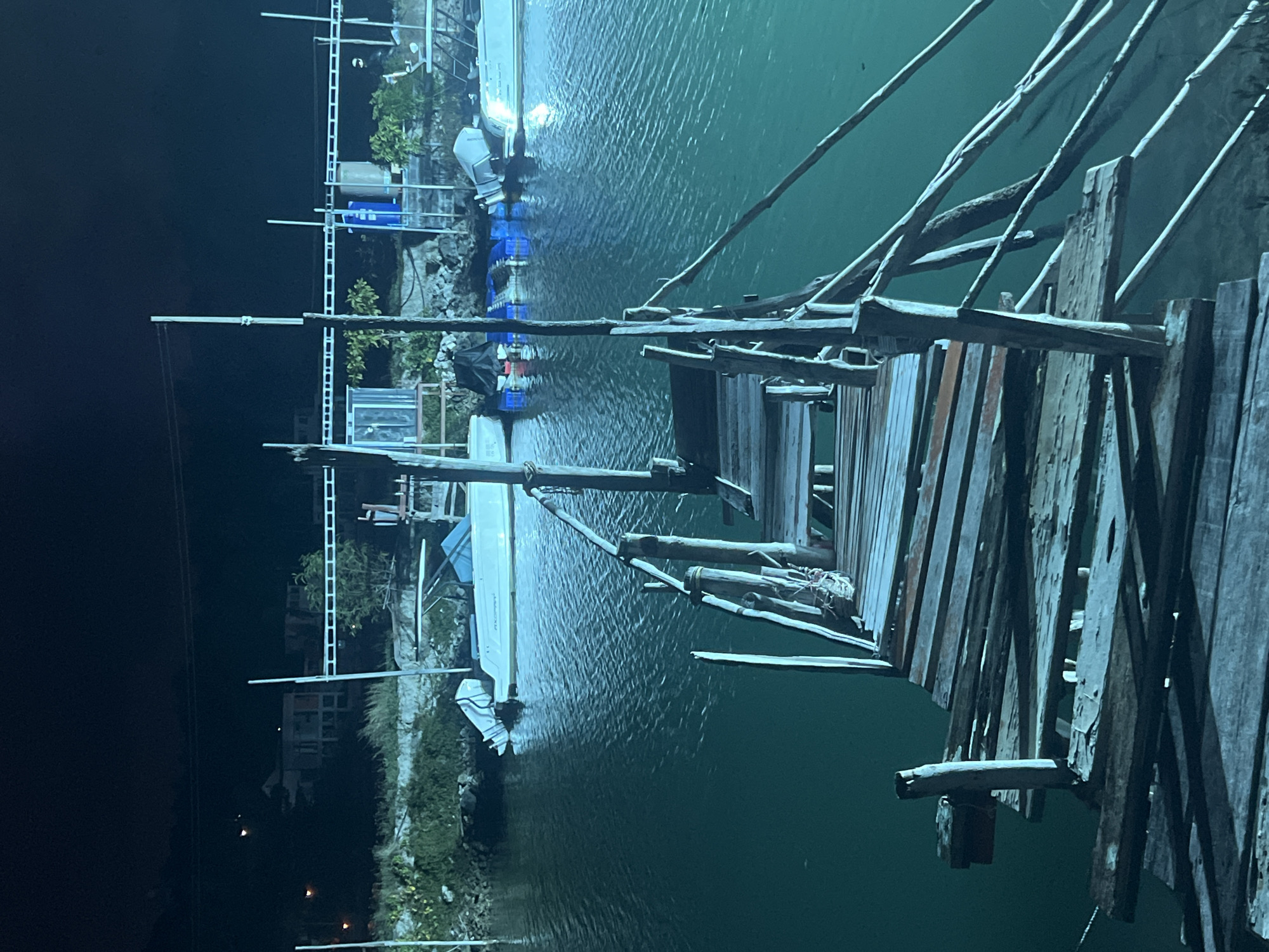 The image shows a nighttime scene at a waterfront location in Ko Pha-ngan District, Thailand. In the foreground, a weathered, wooden pier extends over dark, still water. The pier appears to be in disrepair, with loose planks and a rustic railing made of rough-hewn wood and rope.
In the middle ground, several boats are docked along a shoreline. One white motorboat with an outboard motor is clearly visible, its hull reflecting the ambient light. Other boats and floating structures, possibly part of a marina or docking area, are also present. The water itself is a deep teal color, with ripples indicating slight movement.
The background reveals a low-lying shoreline with vegetation and some buildings. Structures that appear to be houses or possibly small commercial buildings are visible in the distance, illuminated by artificial lights. The overall atmosphere is one of a quiet, possibly remote, coastal area at night, with the emphasis on the rustic pier and the docked boats.