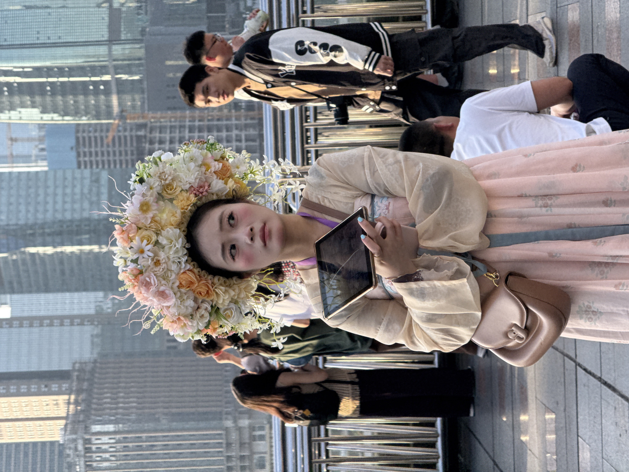 An outdoor, slightly rotated portrait-oriented photograph captures a young East Asian woman dressed in traditional Chinese attire, possibly Hanfu, featuring a light pink patterned skirt and a pale, flowing top. She wears a large, elaborate floral crown composed of white, cream, and peach-colored flowers. Her face, made up with noticeable blush, is turned towards the viewer, displaying a neutral expression. She holds a small, dark tablet with a visibly cracked screen, and her fingernails are painted light blue. A light brown, perhaps leather, shoulder bag hangs from her left shoulder.
The background reveals a bustling urban scene, characterized by towering modern skyscrapers under what appears to be clear daytime skies. Metal railings, suggesting an elevated walkway or bridge, separate the foreground from the distant city view. Several other people are visible behind the main subject. In the upper background, two men stand; one wears a black and white varsity jacket with text that resembles "ESSENTIALS" on the sleeve. Further back and lower, a man in a white t-shirt appears to be resting or lying down. The image seems to capture a moment in a contemporary cityscape where traditional fashion is showcased. The setting is identified as Yuzhong District, China.