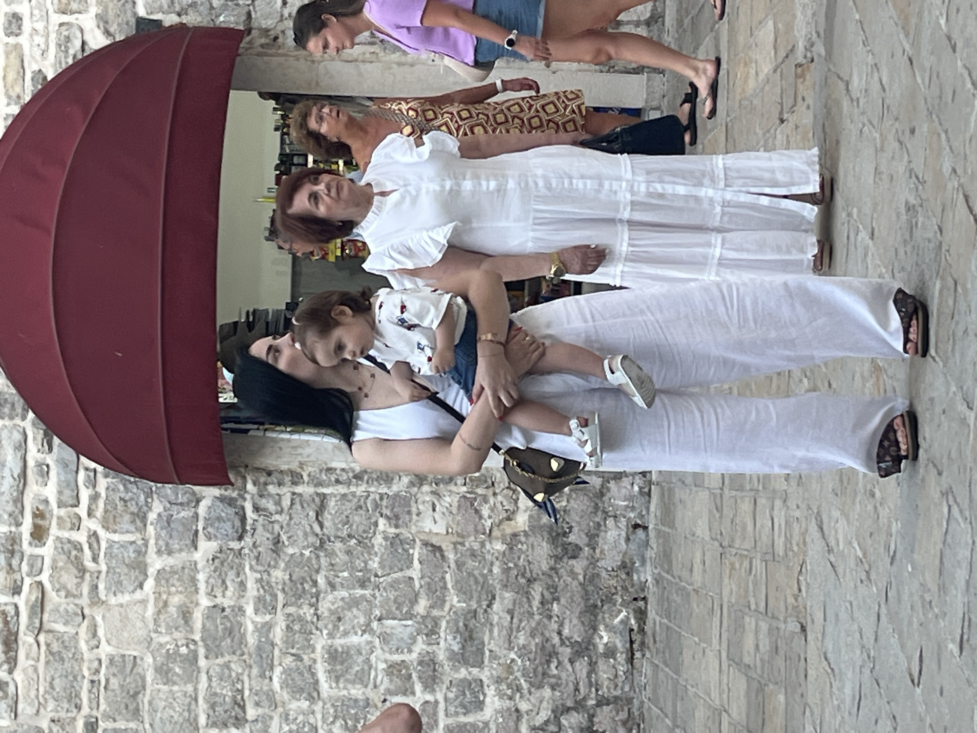 In Budva, Montenegro, a woman in a white linen outfit holds a young child. The child is wearing a white shirt with a colorful print and denim shorts. The woman is also wearing white sandals. Behind them, an older woman in a long white tiered dress with ruffled sleeves stands to the right, also wearing a watch. Further back, another woman in a patterned dress is visible, and to the far right, a woman in a purple top and denim shorts looks towards the right. The backdrop is a stone wall, and a large red awning extends over a shop entrance. The ground is paved with cobblestones.