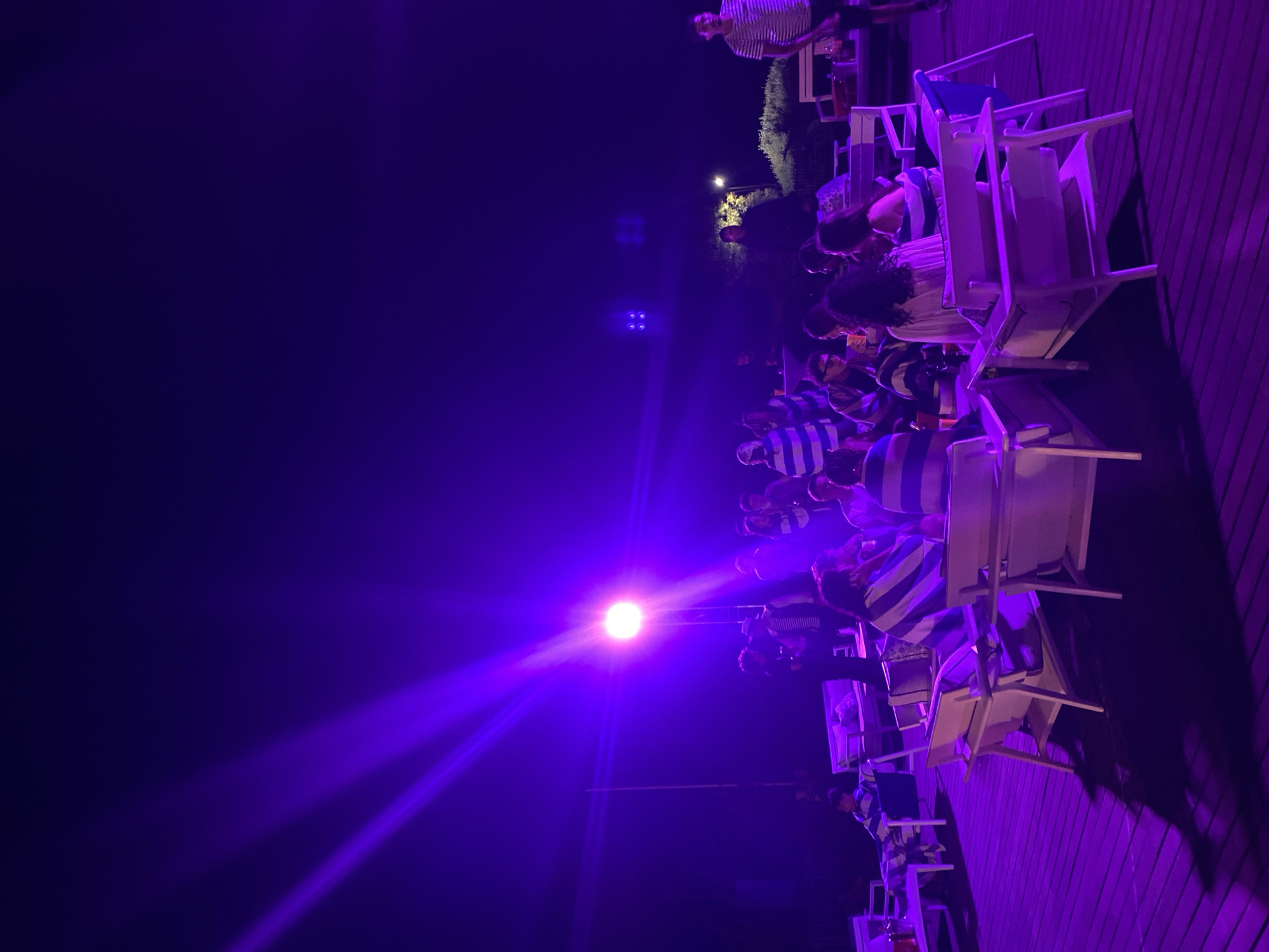 In Taghazout, Morocco, an outdoor gathering is underway at night, illuminated by bright purple lighting. A group of people are seated in white outdoor furniture arranged in a semi-circle on a wooden deck. Some individuals are wearing striped clothing, possibly towels or shirts. In the background, additional people are standing and interacting. A prominent bright light source, casting strong beams, is visible in the upper left quadrant of the image, contributing to the vibrant purple atmosphere. The scene suggests a social event or party taking place in a relaxed, possibly coastal setting.