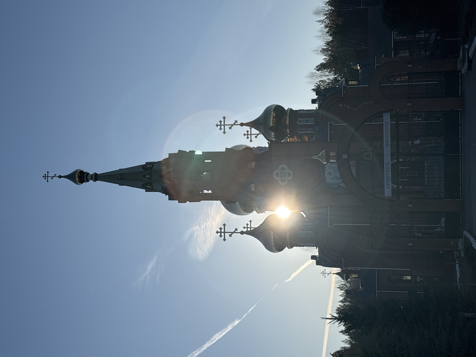The image shows the Exaltation of the Holy Cross Church in Chaikovsky, Russia. The church, with its distinctive onion domes topped with crosses, is constructed of red brick. The sun shines brightly from behind one of the domes, creating a lens flare effect. A gate with a sign in Russian leads to the church grounds. The sky is clear blue with a few contrails from airplanes. Trees are visible in the background. The photograph is oriented sideways.