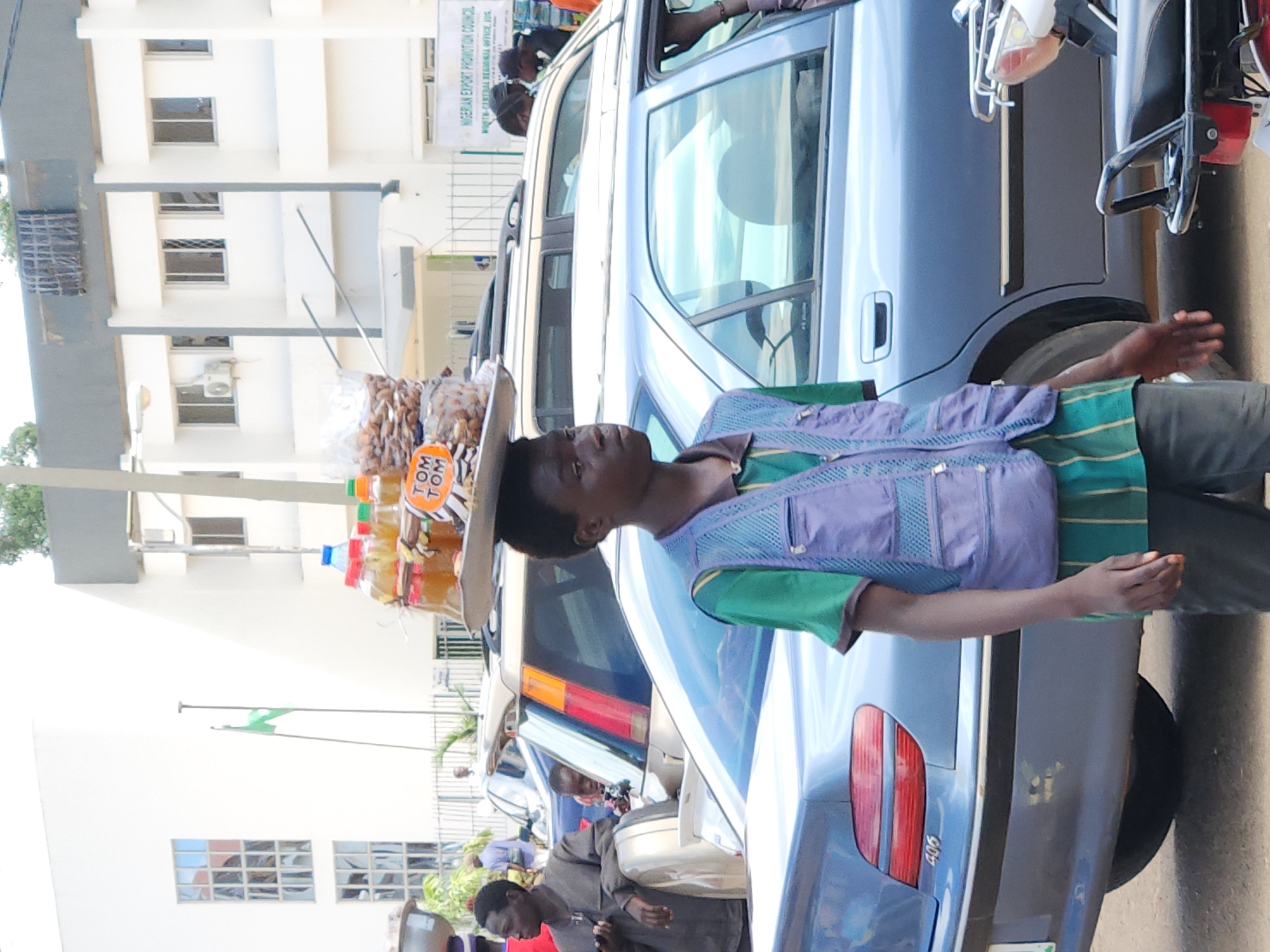 A young man carries a tray of goods balanced on his head in an urban environment. The goods appear to be a collection of bottles and bags of nuts or snacks. He is wearing a green and purple vest over a striped green shirt and dark pants. The background features buildings, several cars, and other pedestrians. One building has a sign that reads "NIGERIAN EXPORT PROMOTION COUNCIL". The scene is outdoors during the day.