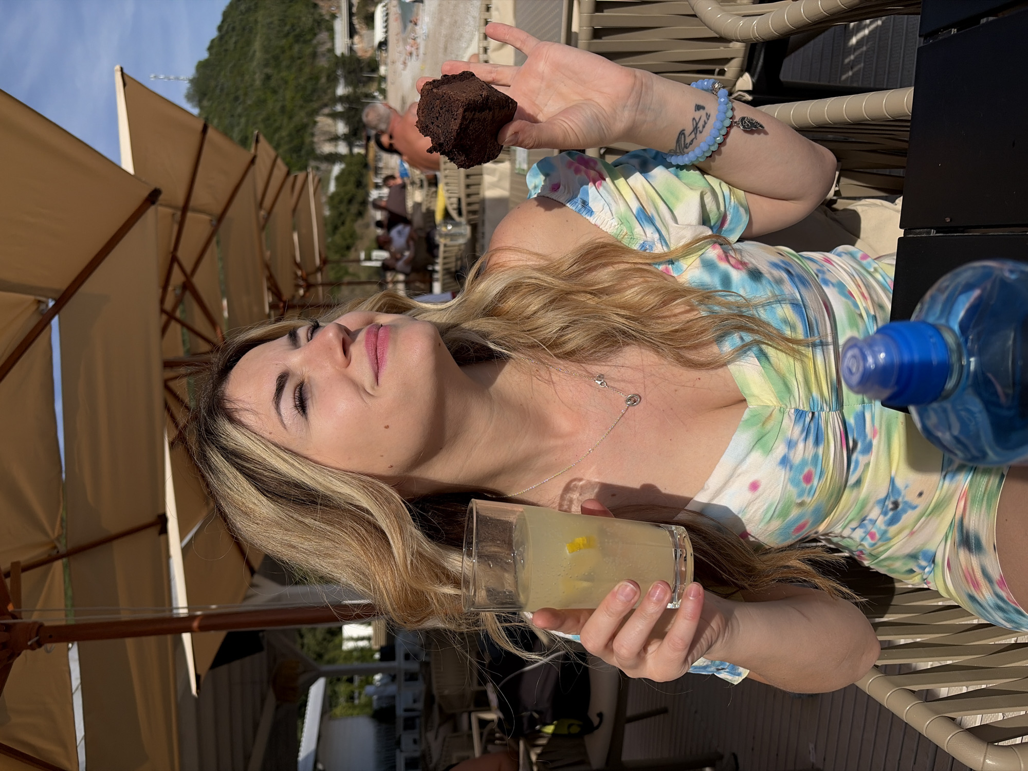 The image features a young woman enjoying a leisurely moment at an outdoor cafe in Budva, Montenegro, under bright daylight. She is seated, looking upwards with a calm and serene expression, under the shade of a large, light brown parasol.
The woman has long, wavy blonde hair with darker roots and is wearing a colorful tie-dye style dress patterned with blue, green, pink, and yellow. She has light makeup, including pink lipstick. In her right hand, she holds a piece of dark, rich chocolate cake, resembling a brownie. Her left hand holds a clear glass filled with what appears to be lemonade, complete with lemon wedges. A delicate silver necklace is visible around her neck, and a light blue beaded bracelet with charms adorns her left wrist. A small, indistinct text tattoo is also visible on her left forearm. A blue plastic water bottle rests on the dark table surface beside her.
The background shows more light brown parasols and beige slatted patio chairs, indicating a cafe or restaurant setting. In the distance, a prominent, verdant mountain or rocky hill rises under a clear blue sky. Blurred figures of other people can be seen in the background. The strong sunlight and shadows suggest it is either late morning or early afternoon.