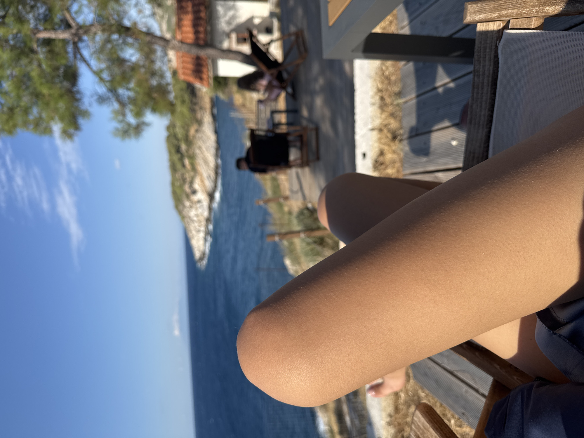 An outdoor scene depicts a person's bare leg in the foreground, positioned across a lounge chair. The leg is tanned and smooth, with a gentle curve at the knee. Behind the leg, a sun-drenched landscape unfolds. The deep blue Aegean Sea stretches towards a distant, rocky coastline dotted with vegetation and a white building with a terracotta roof. A pine tree stands tall on the right, its branches casting shadows. In the middle ground, two other figures are seated in chairs on a wooden deck, facing away from the camera and towards the sea. The overall impression is one of relaxed leisure in a coastal Greek setting.