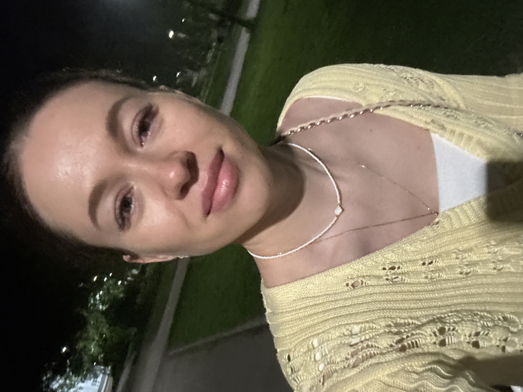 A young adult female is shown in a close-up selfie at night. She has light skin, brown hair pulled back, and is wearing a pale yellow, knitted cardigan over a white top, with two delicate necklaces. The background is blurred and consists of dark areas likely representing the night sky and darker surroundings, with patches of green which are presumably grass. There are also bright blurry spots, likely from distant streetlights or other light sources. Although the city cannot be definitively identified as Perm, Russia, the environment suggests an outdoor location, likely a park or a semi-urban area with some greenery and lighting.