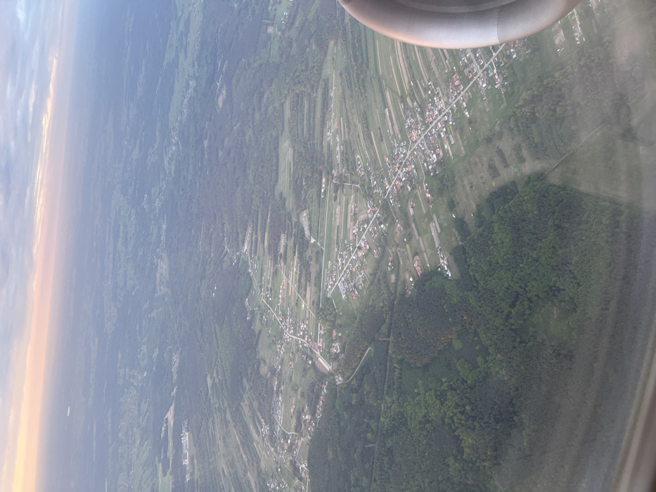 An aerial view of a rural landscape in Jasionka, Poland, captured through the window of an aircraft. The scene is dominated by a large expanse of green forest in the lower left portion of the image. A small village with scattered houses and farms is located in the center-right, stretching out towards the upper right corner. The village features a network of roads and fields, and a few buildings are visible, including a small church and a few houses. The houses are predominantly small and single-story, with roofs of various materials, including red tile and corrugated metal. In the background, distant farmland is visible, with faint traces of trees and buildings, obscured by haze. The sky is a pale blue with a faint pink hue near the horizon, suggesting it may be just after sunrise or before sunset. The image captures a serene and tranquil scene of a rural landscape, with the distant village appearing peaceful and idyllic. The plane's engine cowling, a circular object partially visible on the right edge of the image, is positioned next to the window.