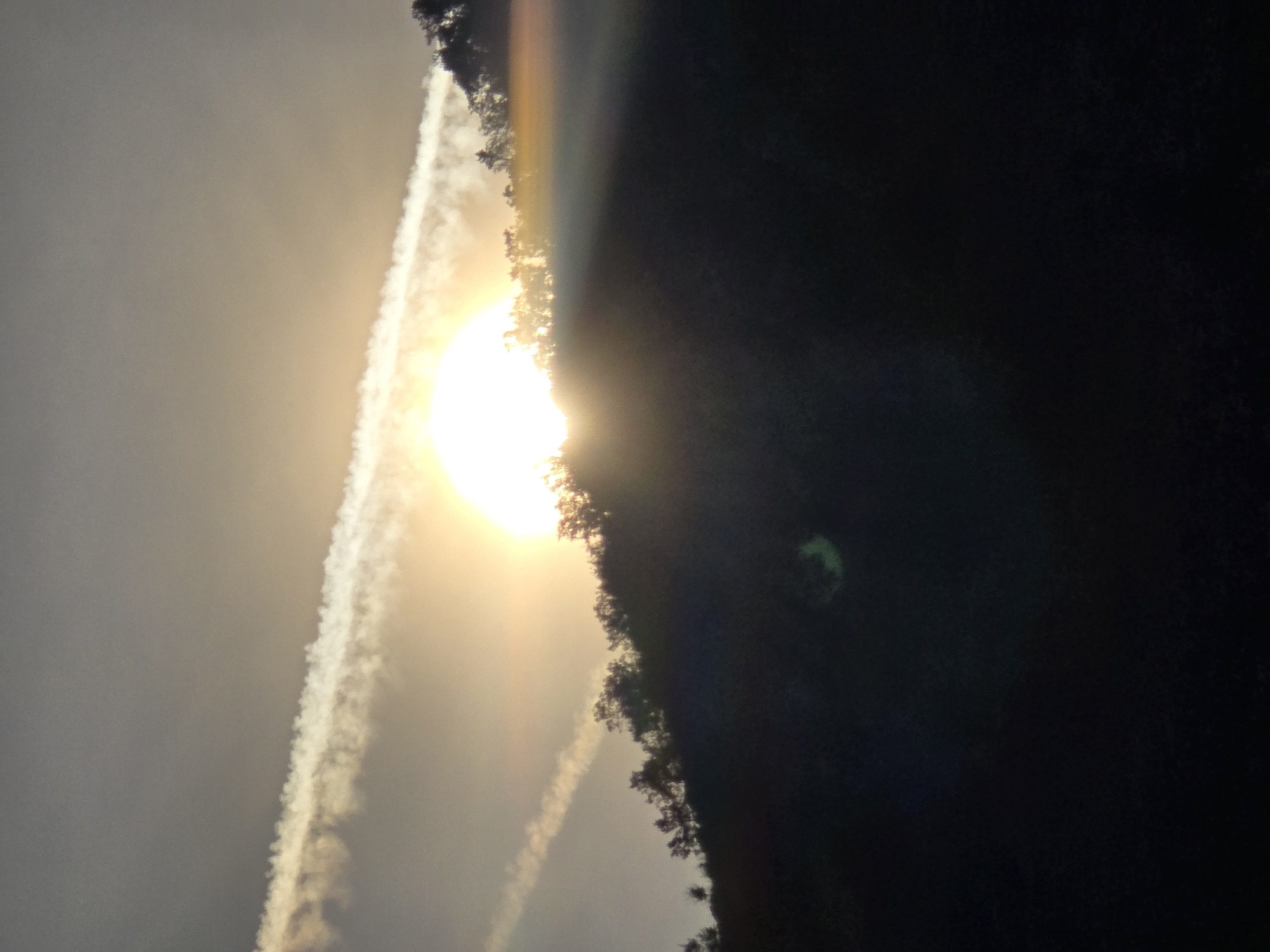 The image depicts a sunset partially obscured by a silhouetted hillside covered in trees. The sun is a bright orb in the sky, casting a warm glow and creating lens flare effects. Two airplane contrails stretch across the hazy sky, intersecting with the sunlight. The lower portion of the image is predominantly dark, suggesting the hillside is casting a shadow over the foreground. The scene appears to be an outdoor landscape, likely a natural setting with vegetation and elevated terrain. The specific location in Podgorica, Montenegro cannot be determined from the visual information provided.