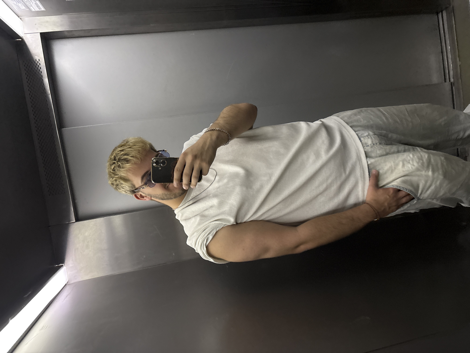 A young man is captured in a mirror selfie, rotated 90 degrees counter-clockwise. He has short, light blonde or light brown hair, a short beard, and is wearing dark sunglasses. His attire consists of a white short-sleeved, textured shirt and light-colored, possibly grey or off-white, relaxed-fit pants. A silver chain bracelet adorns his right wrist, while a smaller, possibly braided, bracelet is visible on his left wrist. He holds a dark-colored smartphone, likely an iPhone, in his right hand, positioned to take the picture. His left hand is casually placed near his left hip.
The setting appears to be a modern interior space, characterized by sleek, reflective, dark grey or silver metallic surfaces, suggesting an elevator or a room with polished, metallic walls. A bright, linear light source, possibly an LED strip, is visible along one of the corners of the space. The overall environment is contemporary and well-lit. There is no discernible text visible in the image. The photo was taken in Sofia, Bulgaria.