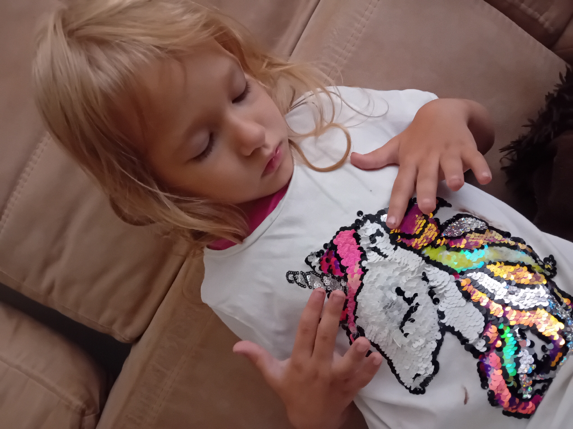 A blonde-haired child wearing a white t-shirt with a sequined unicorn design is sitting on a beige sofa. The child is looking down with closed eyes. Their left hand is resting on the unicorn design on the t-shirt, and their right hand is placed over their chest. The background consists of a blurred beige sofa. The image does not contain information indicating the location in the city of Chuhuiv, Ukraine.