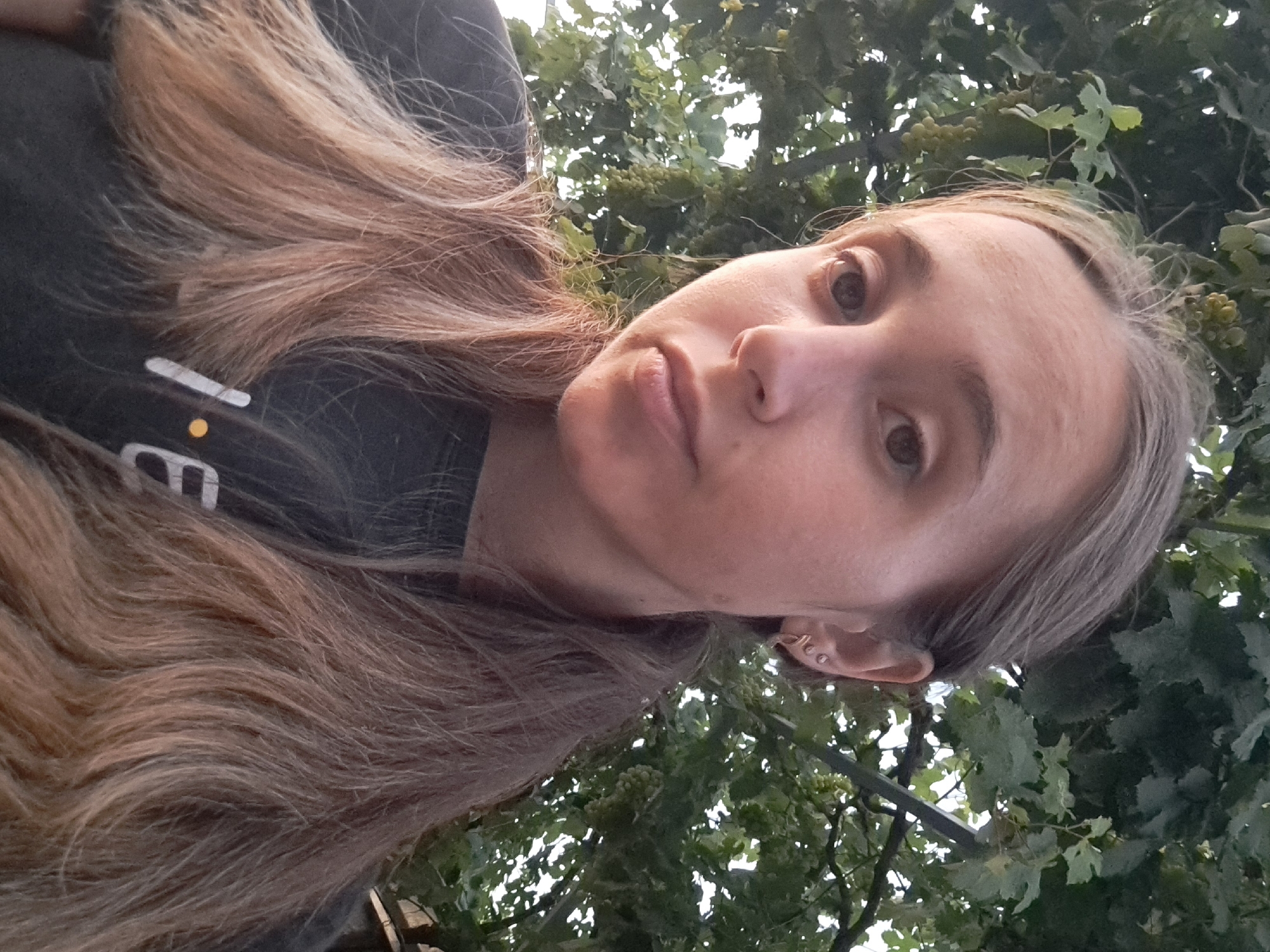 A young woman with blonde hair pulled back into two ponytails stands outdoors in front of a green, leafy background. The woman is wearing a dark gray t-shirt with white lettering and a yellow dot that appears to spell "Friends." Her face is turned slightly to the left, and she is looking directly at the camera. She has light brown eyes and light-colored earrings in her left ear. The background consists of dense green foliage, suggesting a garden or park setting. The lighting indicates it is daytime, possibly late afternoon or early evening. The image was taken in the city of Vypasne, Ukraine.