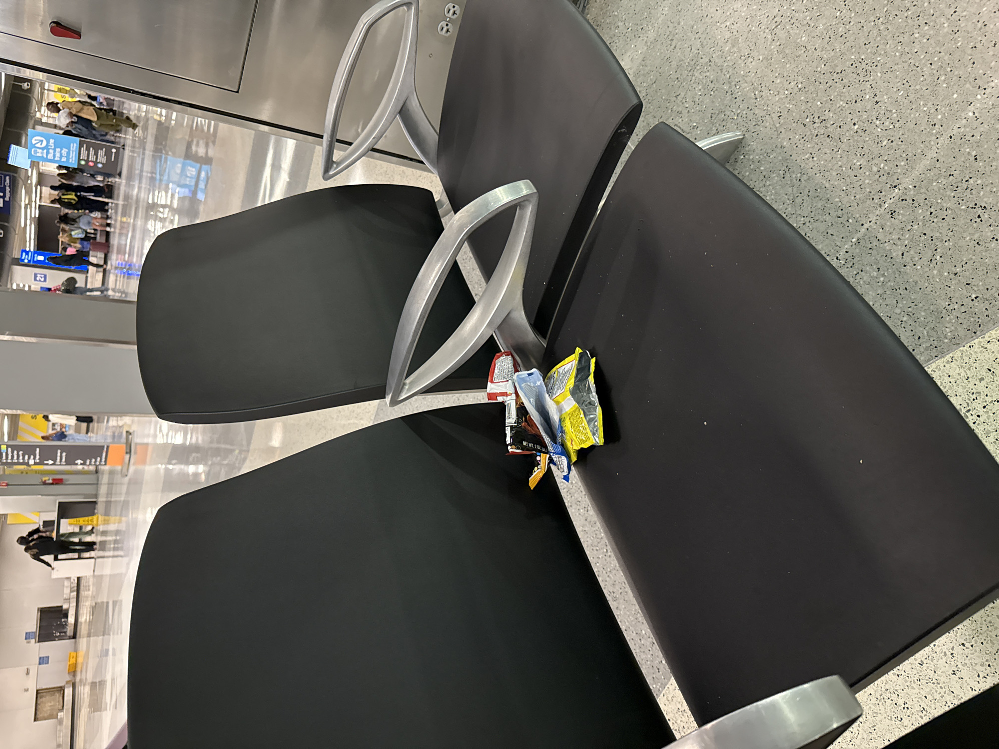 The image shows a row of black airport chairs with silver armrests in what appears to be a waiting area. Litter, consisting of an empty candy wrapper and a snack wrapper, is discarded on one of the chairs. Through the reflective surfaces, glimpses of the airport environment are visible, including signs and the movement of people. Based on the signs visible in the reflection, the image is likely taken at Chicago O'Hare International Airport, in Chicago, United States of America.