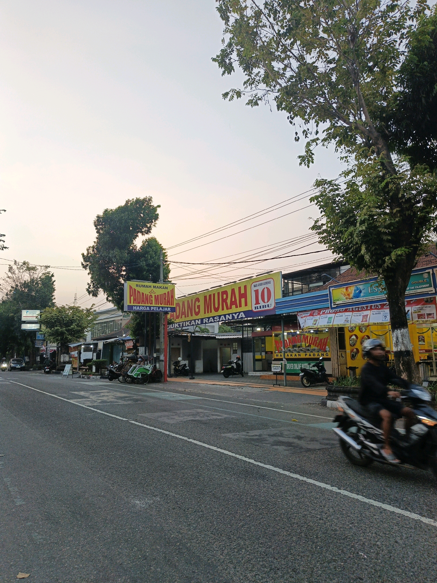 This image depicts a street scene in Klaten, Indonesia, likely in the afternoon or early evening, given the warm, soft lighting. The road is paved and marked with a white line. On the right side of the road, a person is riding a motorcycle, blurred due to motion. Adjacent to the road, there are several businesses, including a restaurant with a prominent yellow sign reading "RUMAH MAKAN PADANG MURAH" (Cheap Padang Food House). Another sign visible next to it says "RUMAH MAKAN PADANG MURAH 10 RIBU" (Cheap Padang Food House 10 Thousand). There are other signs in Indonesian, advertising food and services.
Several parked vehicles, including motorcycles and a green pedicab (becak), are visible on the sidewalk in front of the businesses. Trees line the street, providing shade and greenery. Power lines are strung overhead. In the background, buildings of various architectural styles are present. The sky is overcast with a pale, yellowish hue. A few cars are seen driving in the distance on the left side of the road. The overall impression is of a bustling urban environment, common in Indonesian cities.