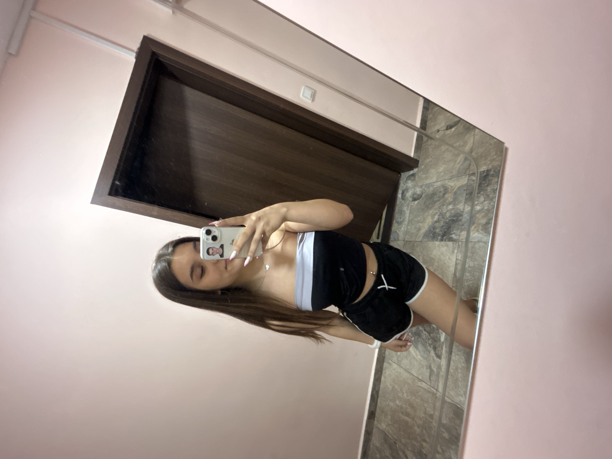 In a dimly lit room with pink walls and tiled flooring, a young woman stands in front of a full-length mirror, taking a selfie with her smartphone. She is wearing a black and grey strapless top and black shorts. The phone, held in her right hand, displays a profile picture of a young man. The mirror reflects the woman, the phone, and a wooden door in the background. The image captures a moment of personal grooming or preparation, possibly within a residential setting in the city of Shumen, Bulgaria.