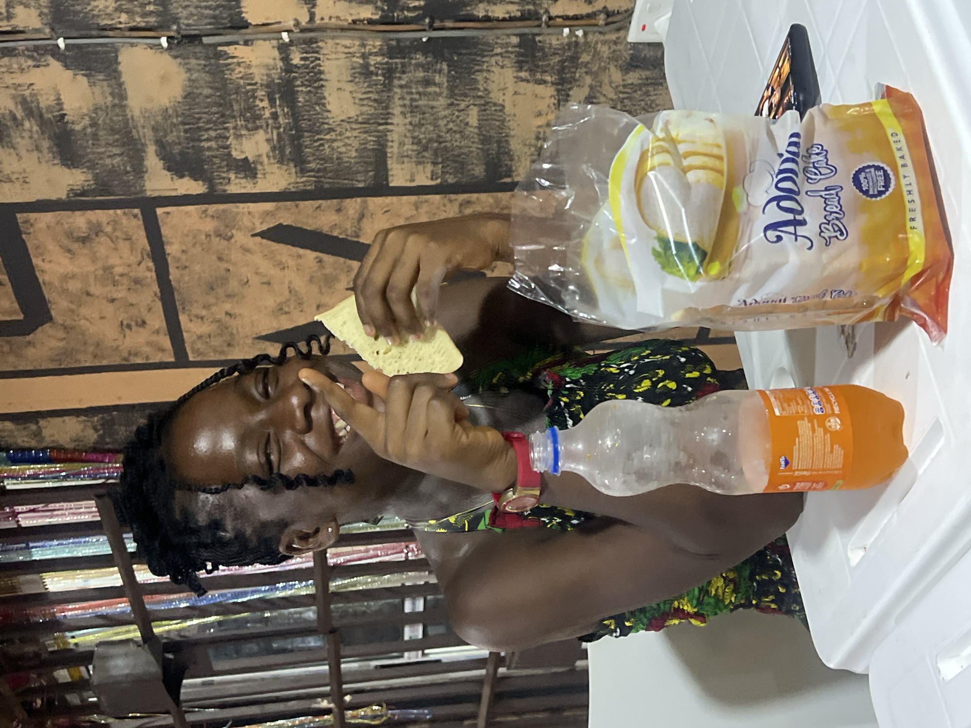 A medium shot captures a young Black woman seated at a white plastic table, seemingly in an outdoor setting or a well-lit indoor space resembling a shop in Nkpor, Nigeria. The woman is smiling broadly, looking to her left, and holding a piece of flatbread in her hands, with her right index finger pointing towards it as if making a point. She wears a colorful patterned dress and a red wristwatch on her left wrist. To her right on the table are two items: an orange-labeled soda bottle, partially filled, and a transparent plastic bag containing a loaf of bread labeled "Adora Bread Cake, Freshly Baked." Behind the woman, the background features a wall with a textured, rustic brown design and vertical elements that appear to be colorful strings or decorations, possibly indicating a vibrant, local marketplace or eatery. The lighting is somewhat dim, casting a warm glow on the scene.