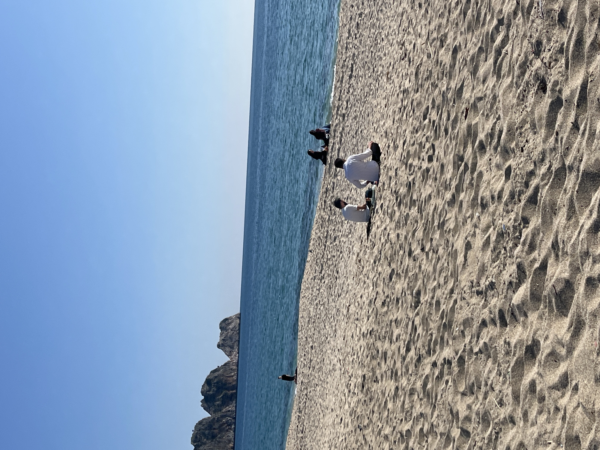 The image displays a sunny coastal scene on a sandy beach in Alanya, Turkey. The foreground and midground are composed of light brown sand with visible ripples and footprints. This sandy expanse meets clear blue-green ocean water that occupies the midground and right background. The upper half of the image is dominated by a clear, unblemished blue sky. In the distant left background, three distinct dark rocky landmasses or small islands rise from the sea.
In the midground, two adult males are seated on the sand with their backs to the camera, facing the ocean. The man closer to the viewer wears a white and blue striped long-sleeved shirt and dark trousers, seated cross-legged. To his left, another man wears a white short-sleeved shirt and dark trousers, also seated, with a small dark object on the sand to his left. Further along the shoreline to the right, two individuals in dark, full-body attire are positioned at the water's edge, either seated or kneeling, with their backs to the camera and partially submerged in the shallow water. To their left, a single individual, similarly clad in dark full-body attire, stands in the shallow sea facing right. No direct interactions between the individuals are observable. The scene suggests a calm, leisurely environment on a clear day.