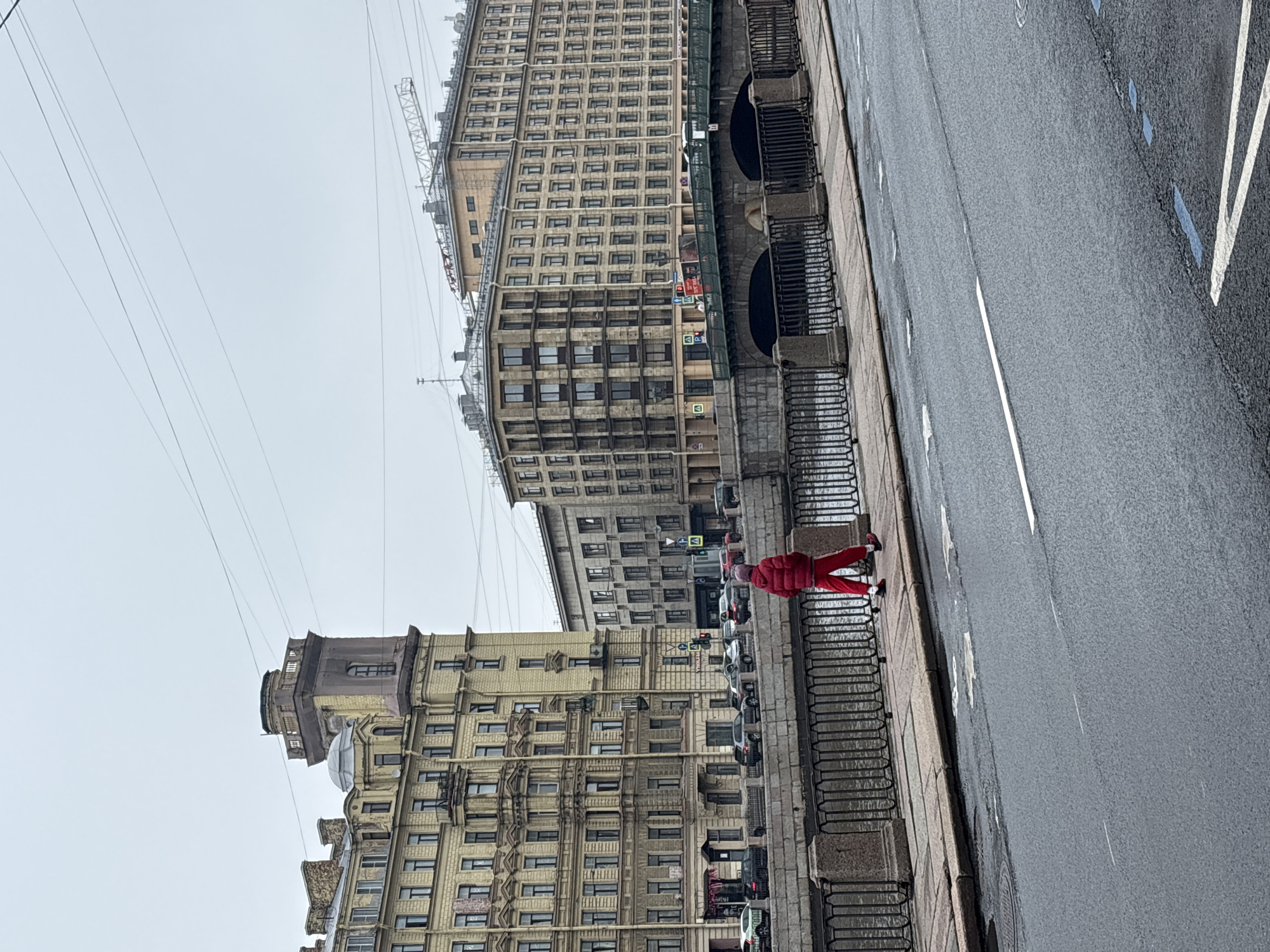 A street scene shows the buildings and canals of Saint Petersburg, Russia. A person wearing red clothing is walking along the sidewalk. Cars are parked along the side of the road. The sky is overcast. Power lines are visible overhead.