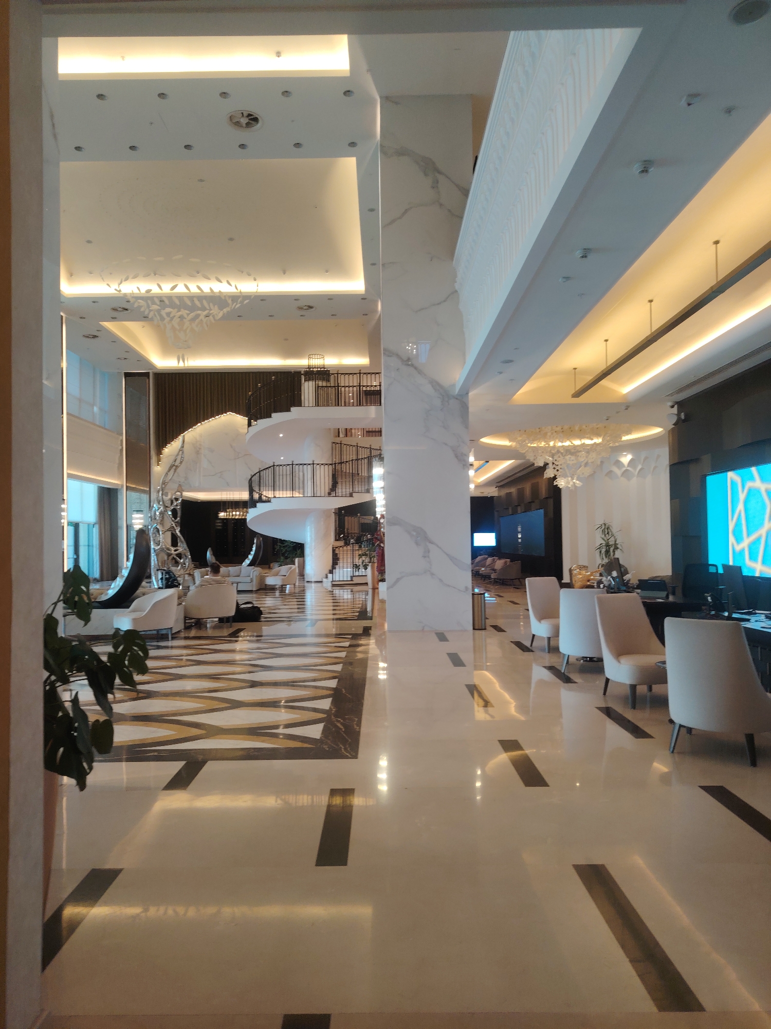 The image depicts the interior of a modern hotel lobby in Konigil, Uzbekistan. The lobby features a high ceiling with recessed lighting and contemporary chandeliers. A prominent spiral staircase with black railings is visible on the left side of the image, leading to an upper level. The floor is polished marble with a geometric pattern. Several white armchairs are arranged in a seating area. A large digital screen displaying a blue geometric pattern is mounted on the wall in the background. A reception desk with computers and chairs is also present. A person is seated in one of the armchairs. The overall aesthetic is sleek and luxurious, with clean lines and a neutral color palette.