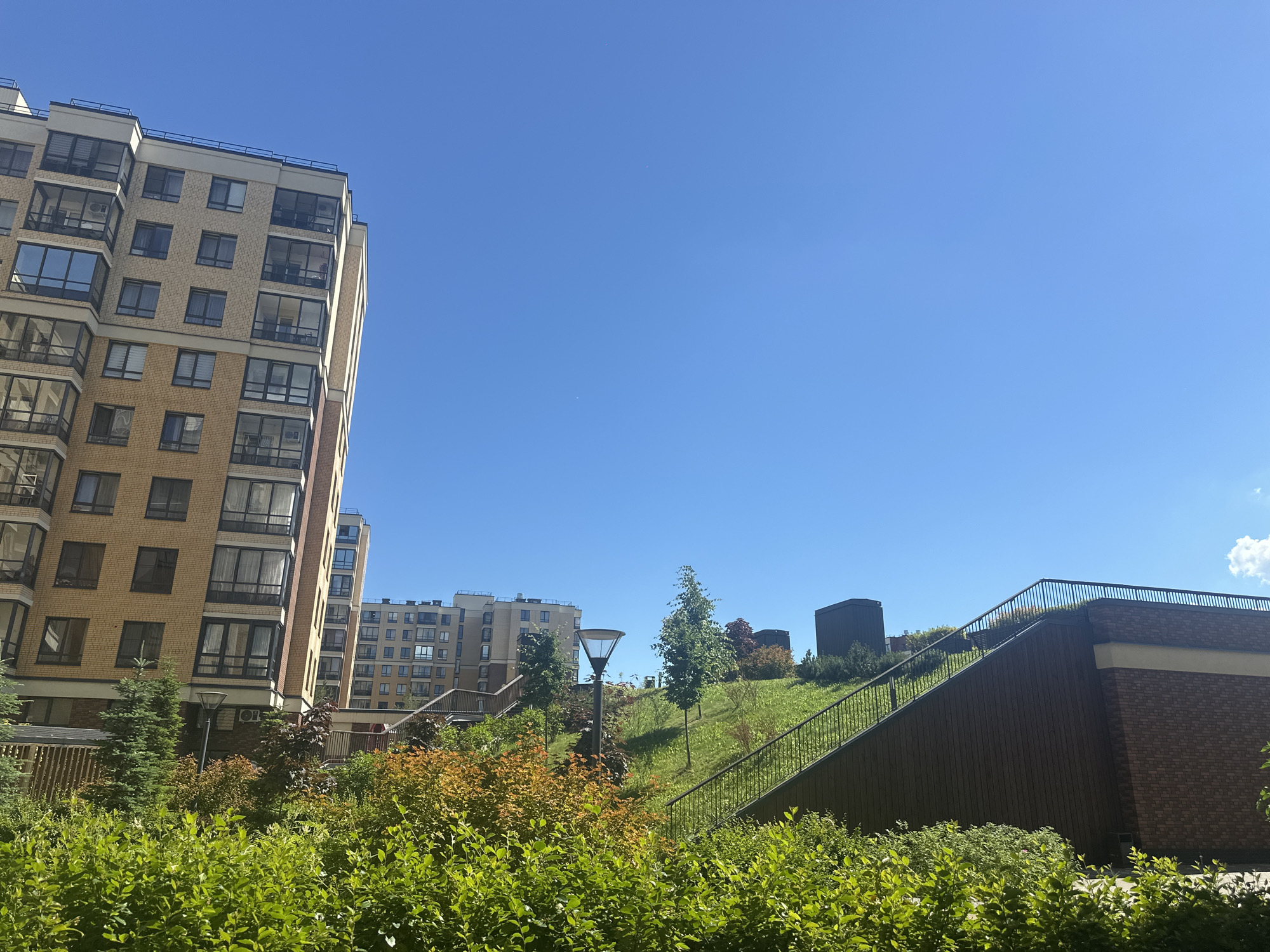 The image shows an exterior view of a modern residential complex under a clear blue sky. On the left side, a tall, multi-story building with beige brickwork and numerous windows is prominently featured. Balconies with glass railings are visible on some floors. In the foreground, lush green bushes and some trees with autumn foliage create a vibrant natural element. Behind the foliage, a sloped grassy area with pathways and staircases is visible, leading up to a dark-colored structure on the right. A lamppost stands near the center of the frame, amidst the greenery. Several other residential buildings of varying heights and designs are scattered in the background. The scene suggests a well-maintained urban environment with a focus on green spaces.