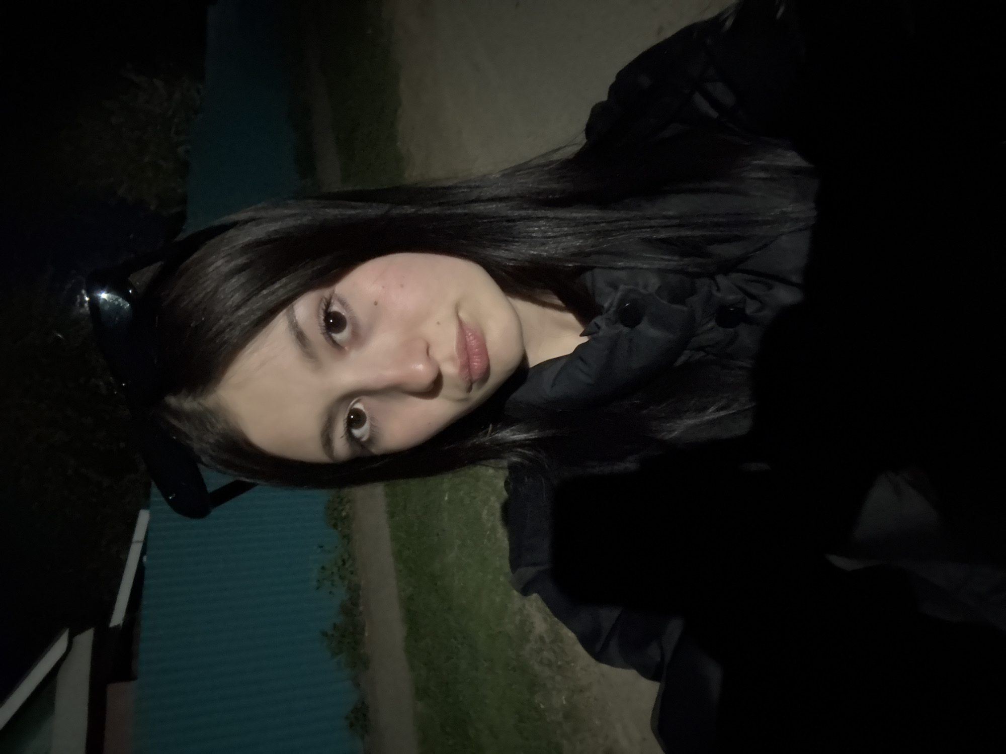 A young woman with dark hair and light skin is posing for a selfie in an outdoor location, possibly in the city of Фоки, Russia, as indicated in the instructions. She is wearing a dark-colored jacket or coat. The background is dark, but reveals some green foliage and what appears to be a road or path. There is also a glimpse of a light-colored wall or structure on the left side. It appears to be nighttime, based on the low lighting conditions. The woman is looking at the camera with a slight smile. A pair of sunglasses can be seen in the background.