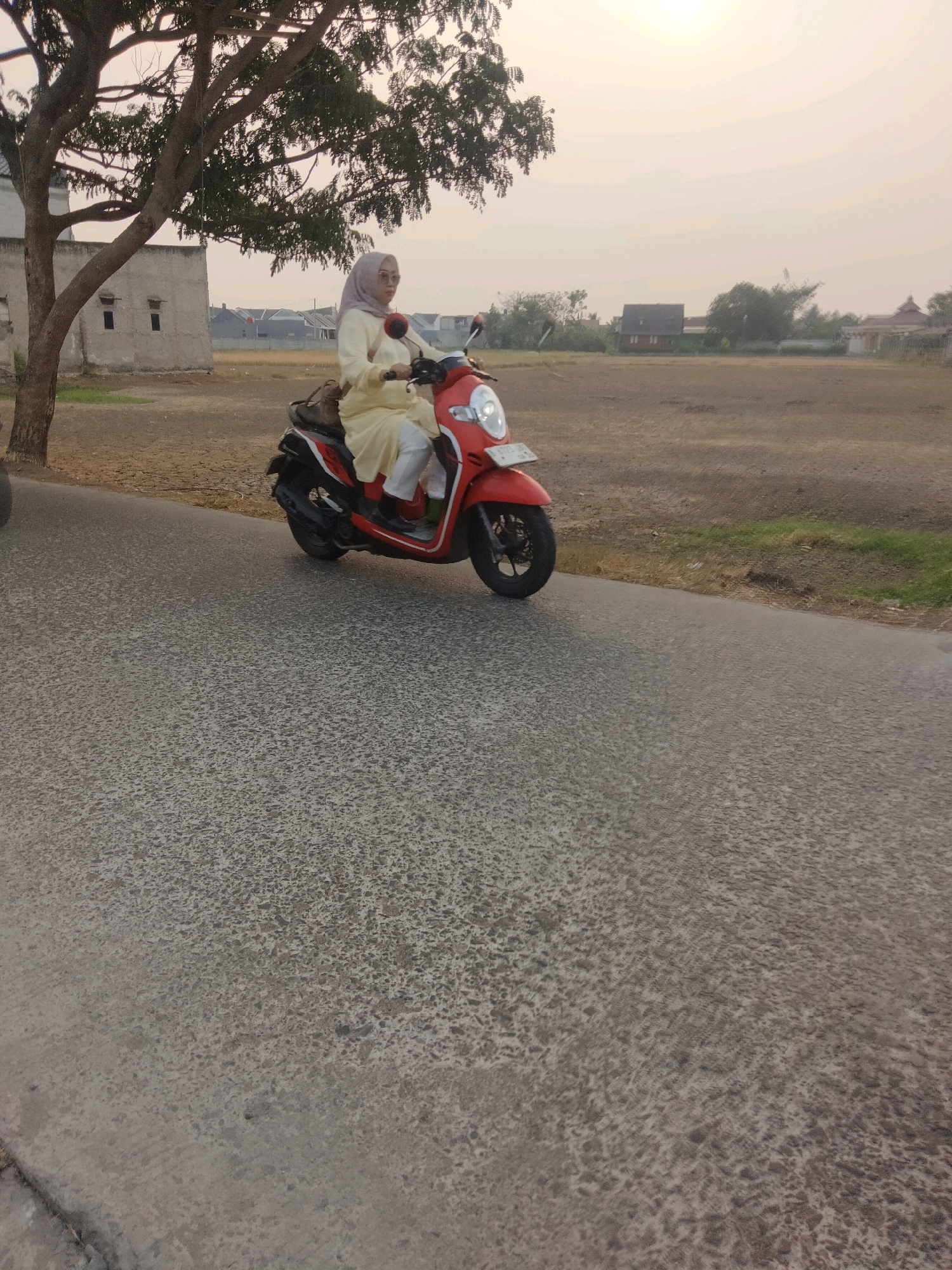 A woman wearing a light yellow hijab and matching outfit is riding a red and black scooter on a paved road in Suka Sari, Indonesia. She is looking forward as she rides. The road surface is textured with dark grey and lighter grey patches. To the left of the road is a patch of dry, brown ground with a large tree with green leaves and a portion of a light-colored building. Behind the tree and building, further houses are visible, with red-tiled roofs. To the right of the road is a large, open, flat field of dry, brown dirt with some sparse green vegetation along the edge of the road. The sky is hazy and a pale yellow, suggesting it is either early morning or late afternoon. Several other buildings and trees are visible in the distance. The overall environment appears to be a semi-rural or suburban area.