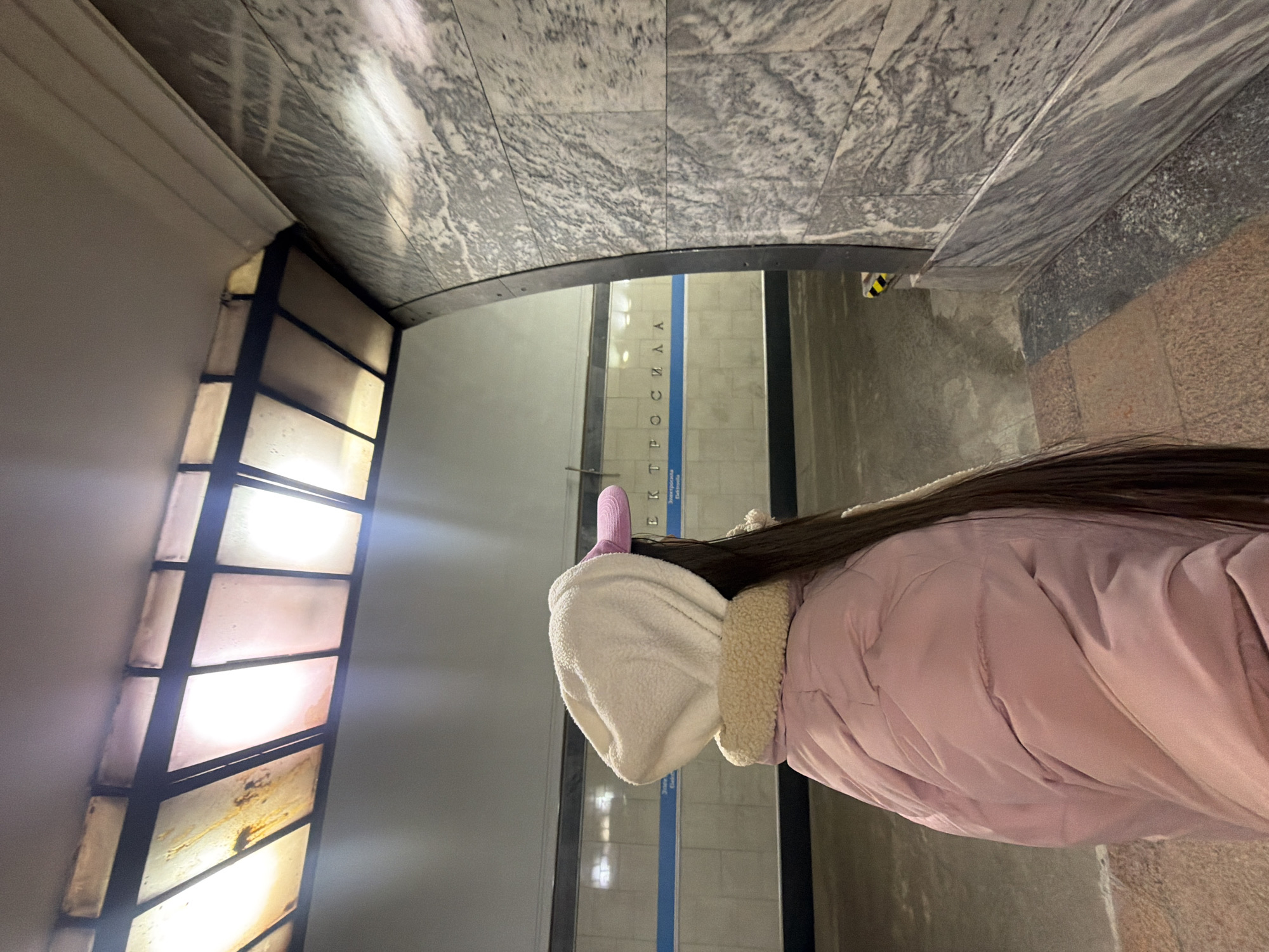 A woman in a pink winter coat with a white fur-lined hood and pink bunny ears stands in a Saint Petersburg metro station in Russia. The station walls are lined with grey and white marble. The text "ТРОСИЦКАЯ" is visible on a sign. Behind the woman are light boxes set in black frames. The ground appears to be tile.