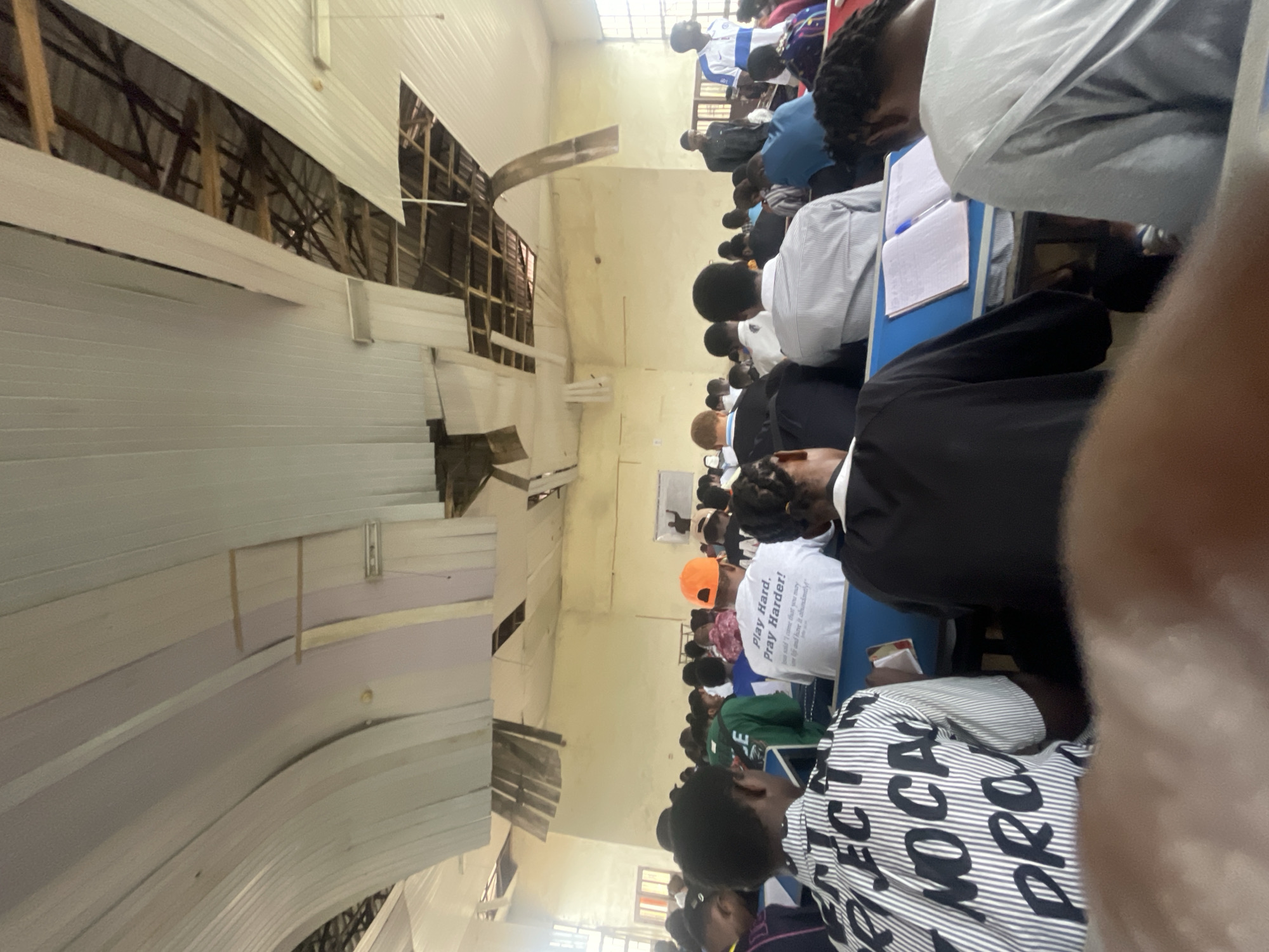 In Ihiagwa, Nigeria, an indoor gathering of people is taking place in a large room. The ceiling is damaged, with sections of the white paneling pulled away, revealing exposed wooden beams and metal supports. The walls are painted a pale yellow. In the foreground, the backs of several individuals are visible, seated at blue tables. One person in the center wears a white t-shirt with the text "Play Hard. Pray Harder!" printed on it. The rest of the attendees are facing forward, some looking at notebooks and pens on their tables. A whiteboard is visible on the far wall, with a figure standing in front of it. The room appears to be a lecture hall or a meeting space.