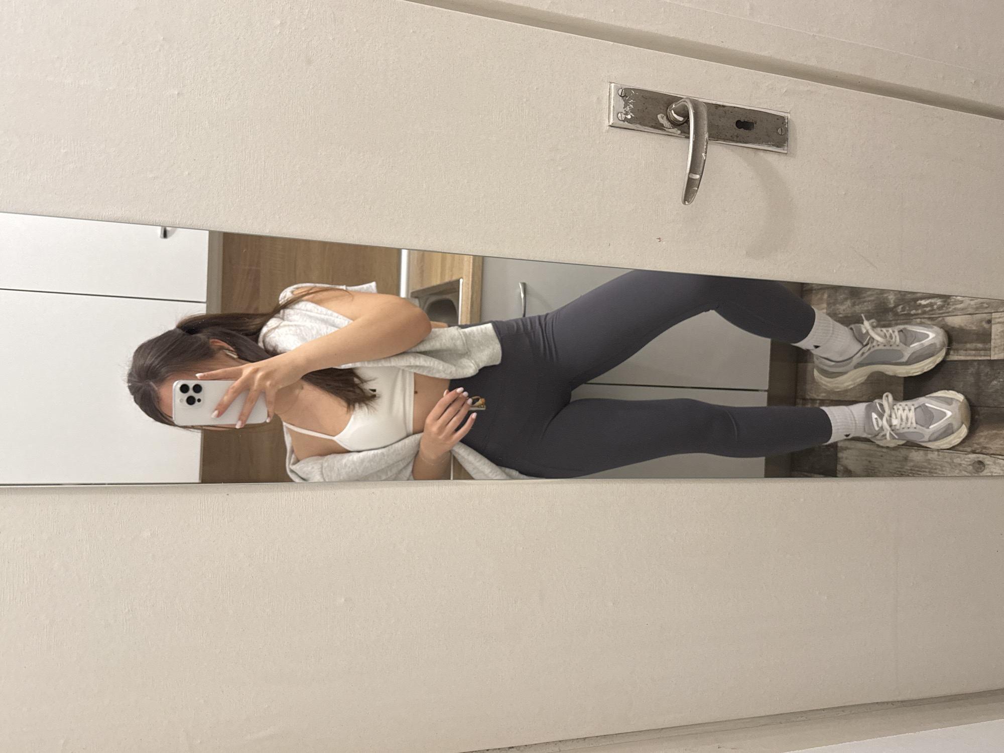 A woman is reflected in a bathroom mirror, holding a smartphone in front of her face. She is wearing grey leggings, a white sports bra, and a grey sweatshirt. She stands on grey and brown floor tiles. The bathroom has white cabinets, a silver faucet, and a white door with a silver handle. The location cannot be determined from the image alone.