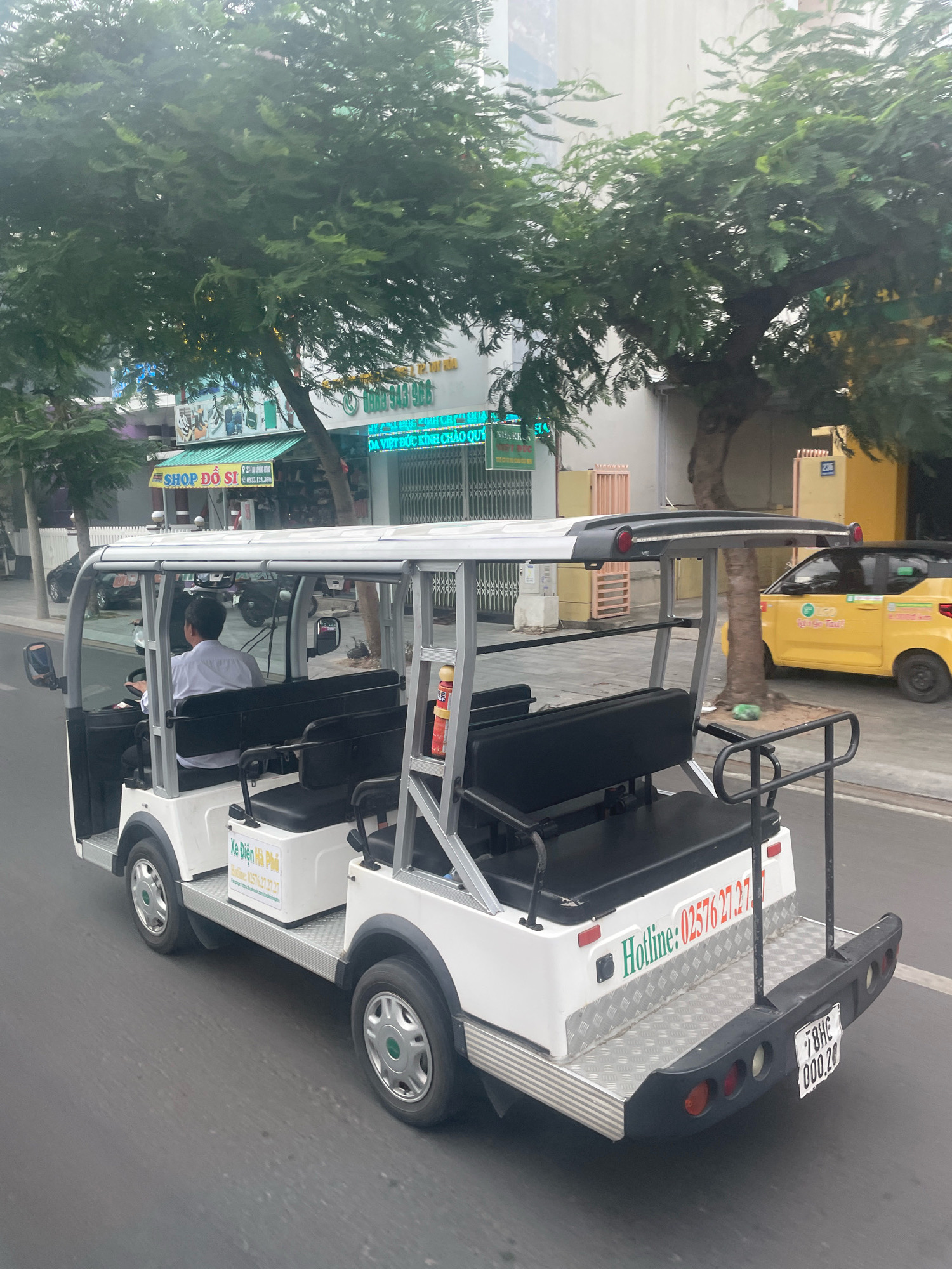 The image shows a white electric cart with a black roof and seating. A man wearing a light blue shirt is driving the cart. The cart has the text "Xe Điện Hà Phú" and a hotline number "02576 27.27.27" on its side. The cart is on a paved road in front of buildings with signs in Vietnamese, suggesting it is in Vietnam. One sign reads "SHOP ĐỒ SỈ" and another shows a phone number. A yellow taxi is visible to the right of the cart. The license plate on the back of the cart reads "78HC 000.20". The overall scene depicts street life in Tuy Hoa, Vietnam.