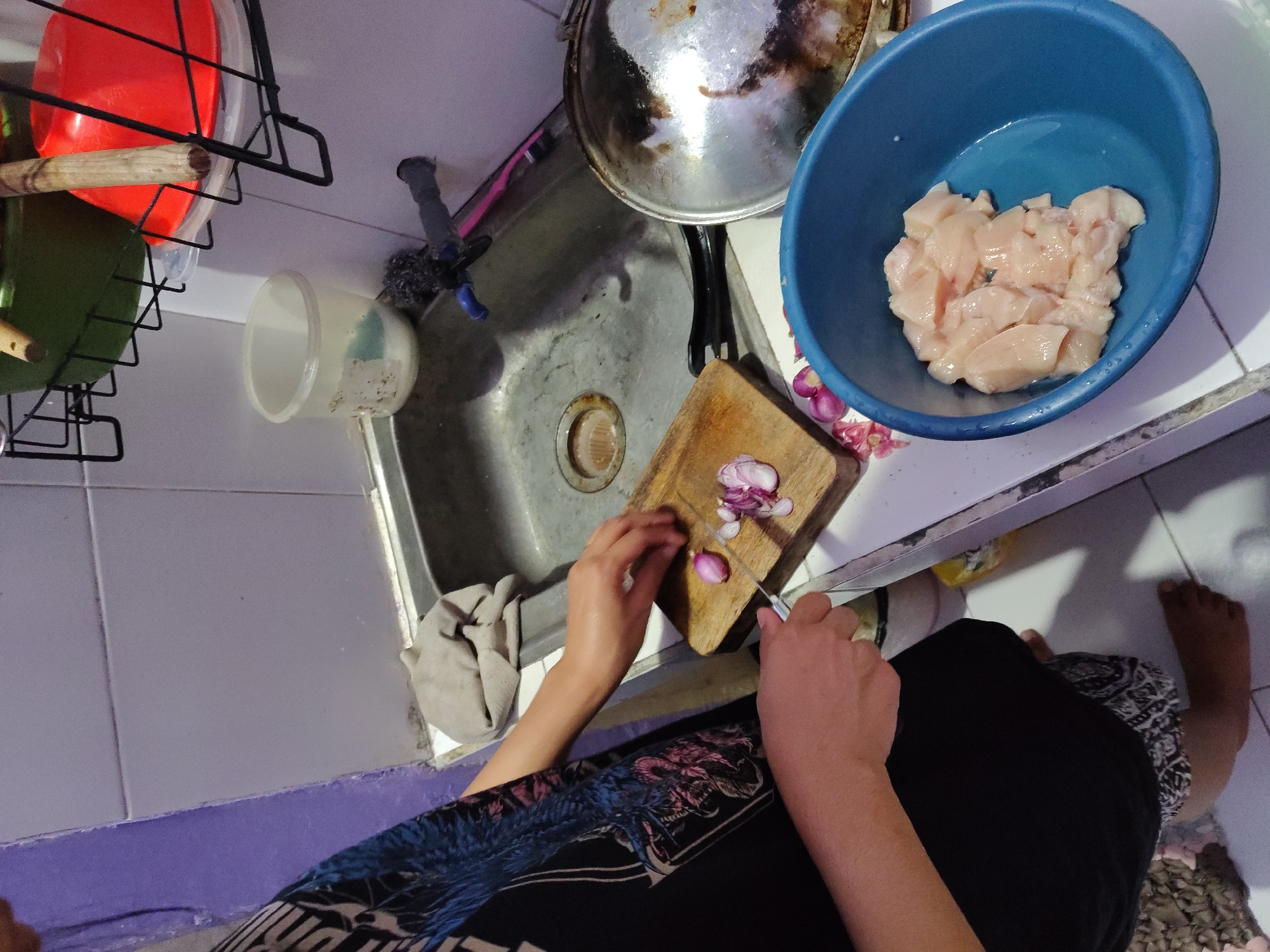 This is a close-up, medium-angle shot of a person in a kitchen in Surabaya, Indonesia, preparing food. The person, seen from the chest down, is wearing a black shirt with a colorful, patterned design and dark pants. They are standing in front of a stainless steel sink. The person's hands are actively engaged in chopping red onions on a small wooden cutting board. A small knife is also visible on the board, along with several sliced onion pieces. To the right of the cutting board, a pile of chopped red onions is scattered on the countertop. In the foreground, a bright blue plastic bowl contains several pieces of raw, cubed chicken. The kitchen environment appears utilitarian, with a tiled backsplash and various kitchen items visible in the background, including a faucet, a sponge, and a pan with a lid.