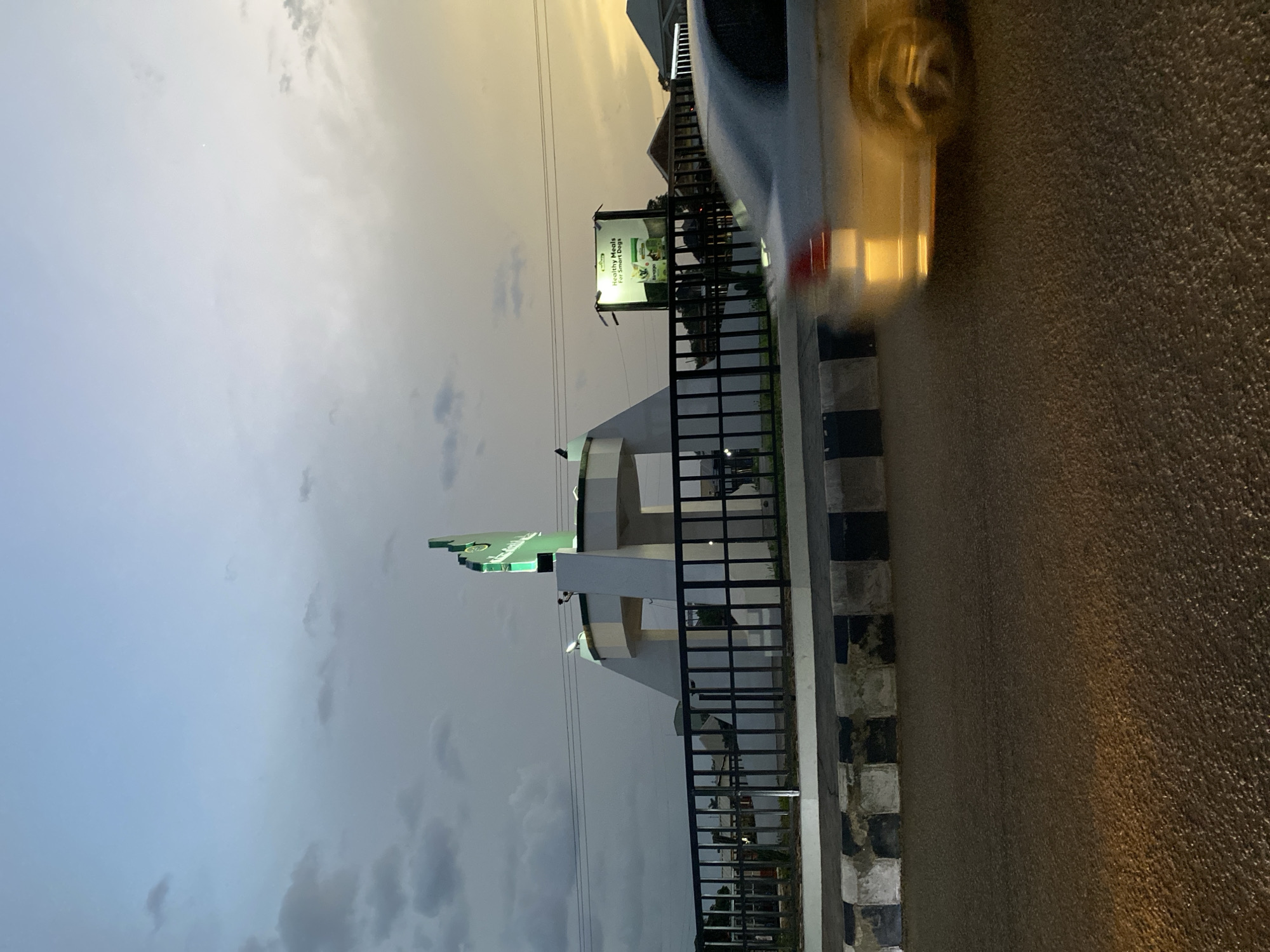 The image shows a view from a vehicle in Kuru Dungus, Nigeria. The foreground includes part of a car, with a blurred headlight. Along the roadside is a black metal fence and a curb with alternating black and white tiles. Behind the fence, there is a white structure with a green map of Nigeria on top. A billboard is visible in the background, displaying a "Healthy Meals for Smart Dogs" advertisement. The sky is overcast with grey and white clouds. The time of day appears to be evening.