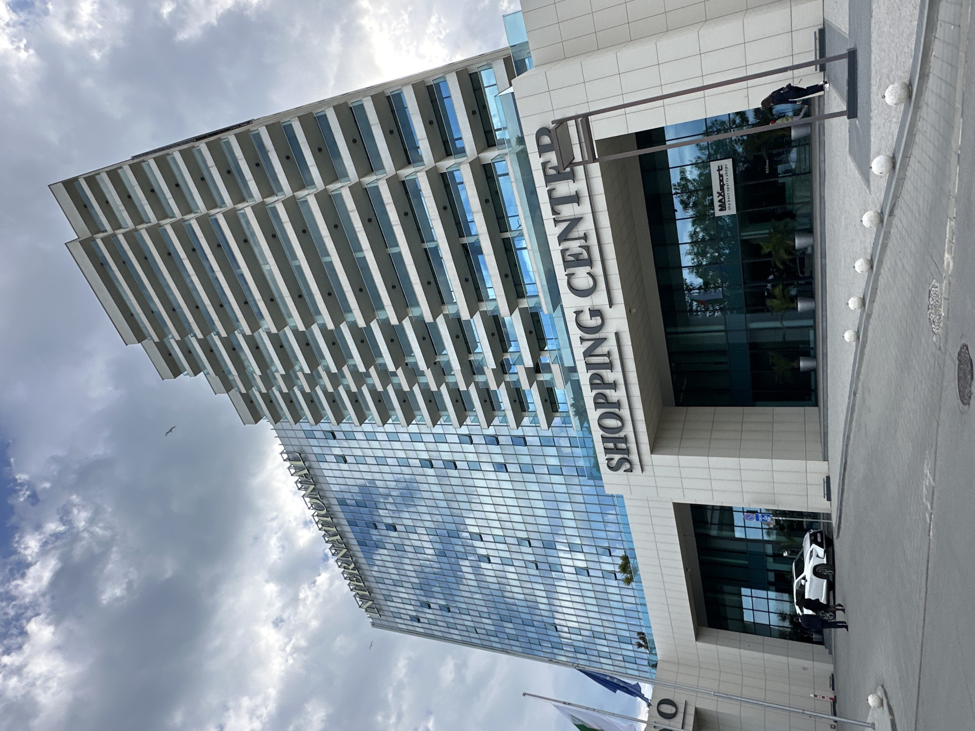 The image shows the Grand Mall Varna shopping center in Varna, Bulgaria. The building is a tall, modern structure with a glass facade and white balconies. A sign on the building reads "SHOPPING CENTER" vertically. At the entrance, two people are interacting near a white car. Flags are displayed in front of the building. The sky is cloudy. A pedestrian is walking by the entrance of the shopping center.